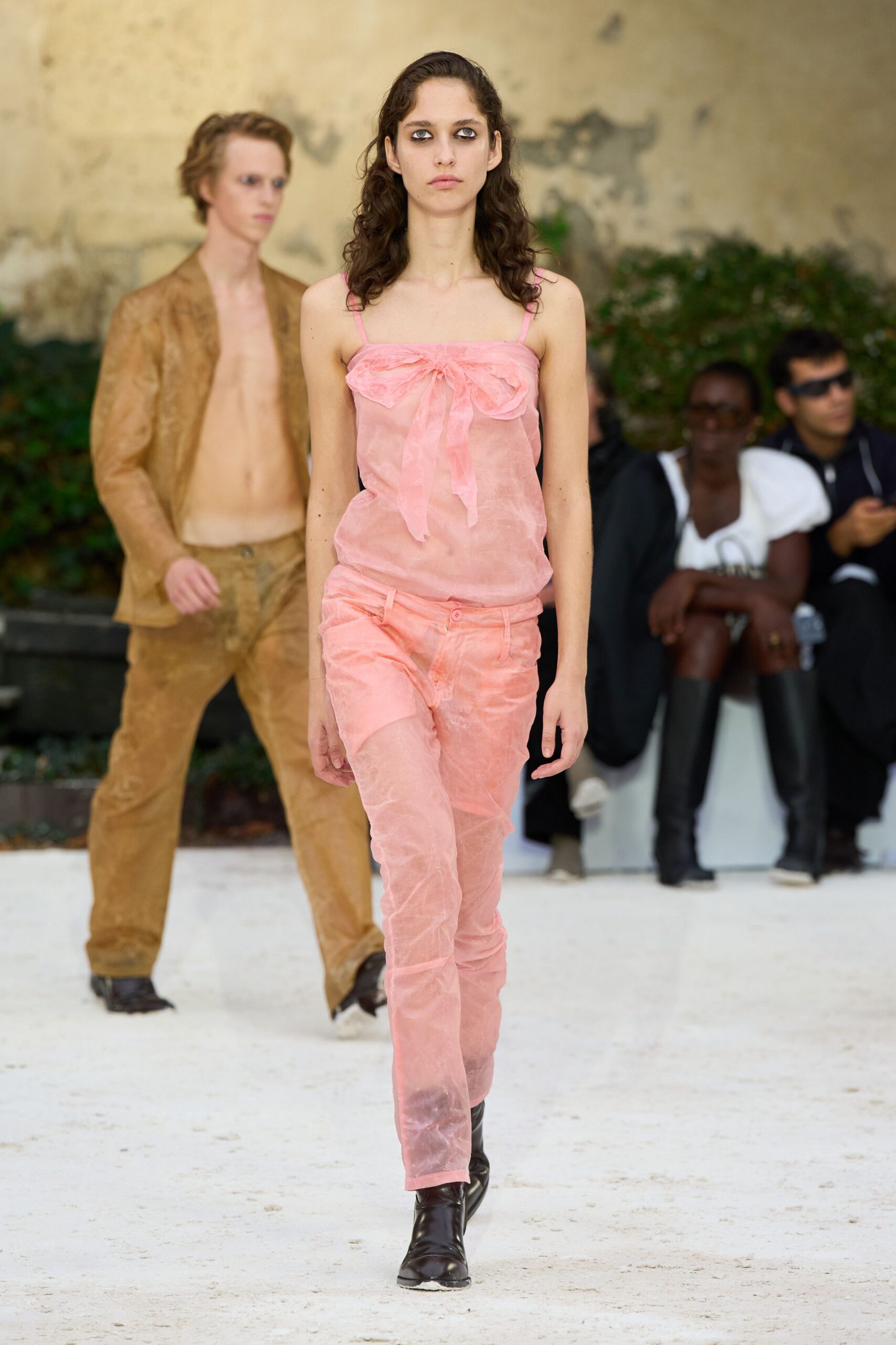 Sheer blush camisole with a gauzy bow knotted across the bust. Matching translucent trousers over short pink underlayer, single button at the waist. Black square-toe ankle boots. Walking across a dusty courtyard before the heat breaks.