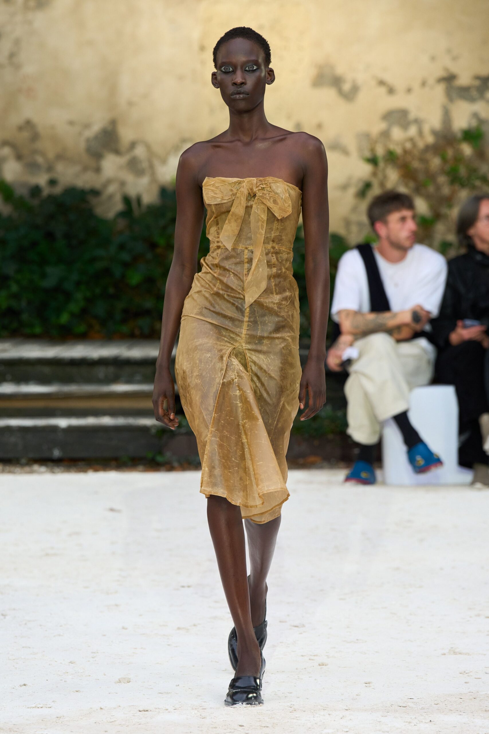 Strapless ochre sheer dress, crushed and ruched with a drooping bow at the bust. Black patent flats with square toes. Crossing a sunburnt courtyard before the wedding guests sit down.