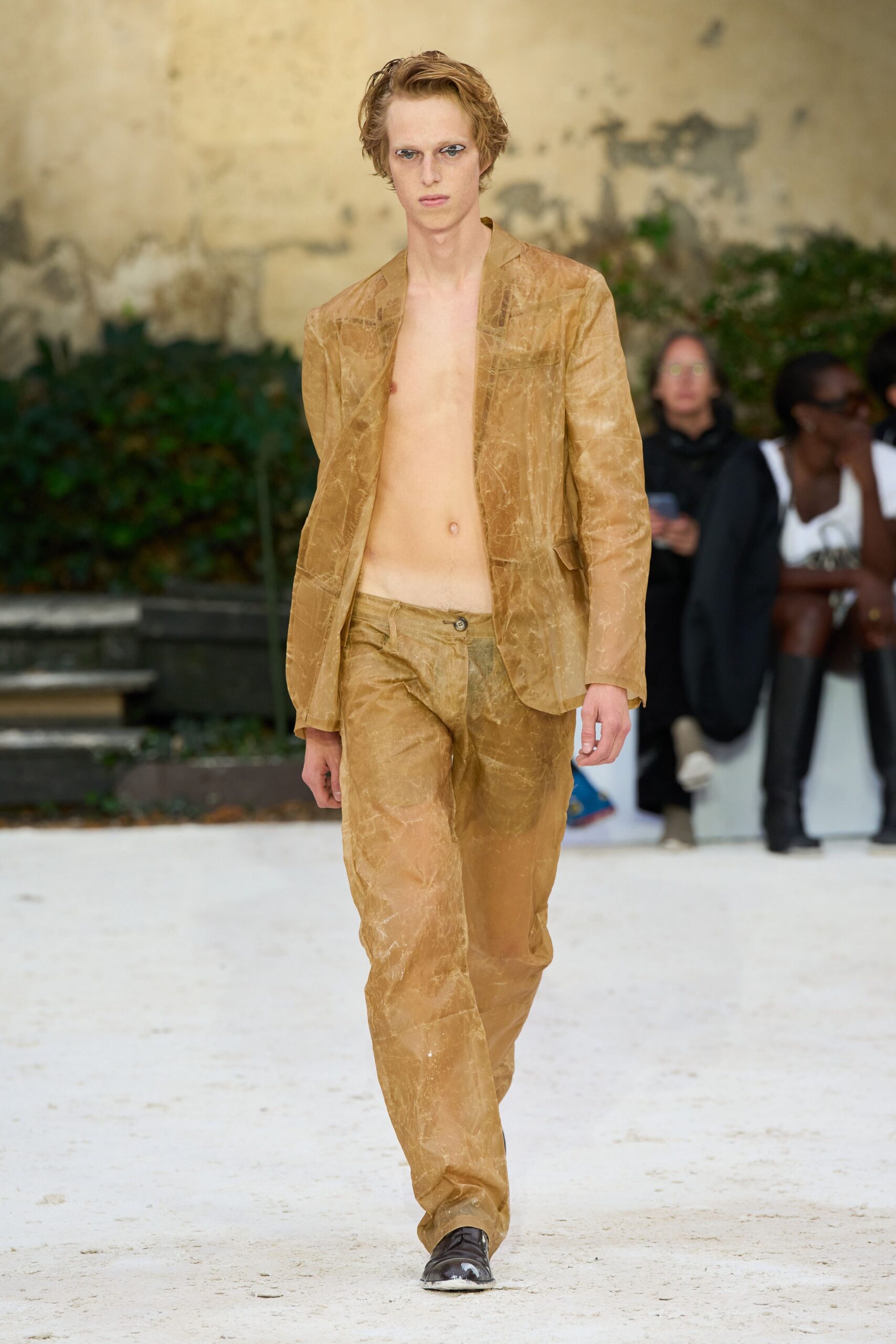 Camel blazer in crinkled translucent fabric, worn bare with low flap pockets. Matching straight trousers, slightly dropped waist and a single dark button. Black leather square-toe shoes, chalked white at the tip. Crossing a gravel courtyard before noon, jacket already sticking to the skin.