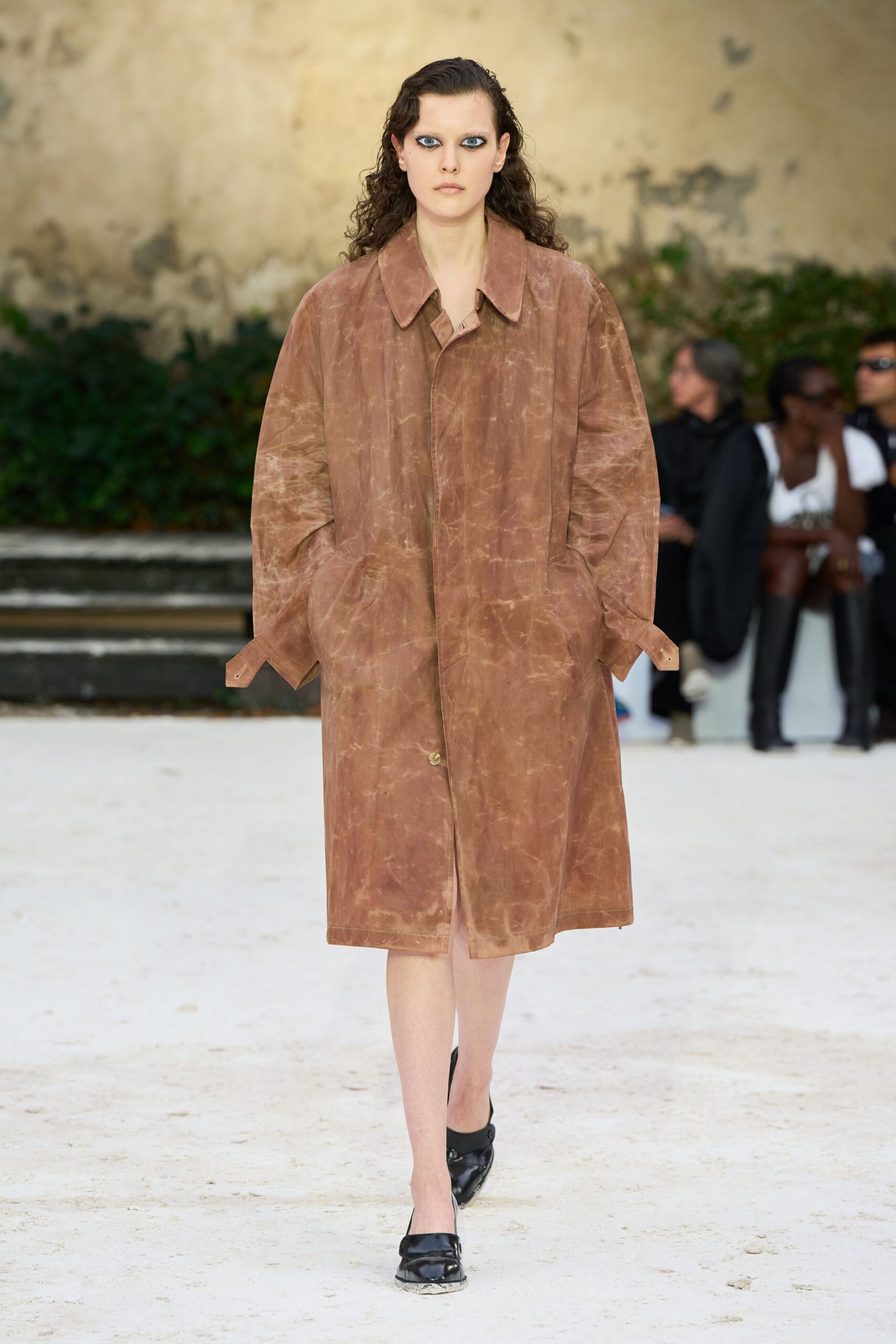 Dusty russet leather coat, marbled with pale abrasion lines and strap cuffs. Blush slip of a dress peeking below the placket. Black leather mules with square toes and a scraped front edge. Crossing a gravel courtyard after the storm dried into chalk.