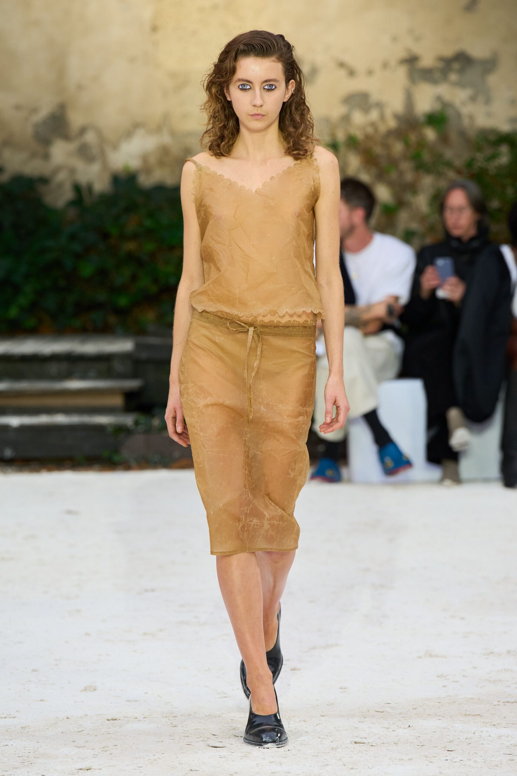Sheer camel camisole with scalloped lace edging and a crumpled finish. Matching pencil skirt in cloudy translucent fabric, tied with a narrow drawstring. Black pumps, square at the toe. Walking across a sunstruck courtyard after swimming, still half dressed.
