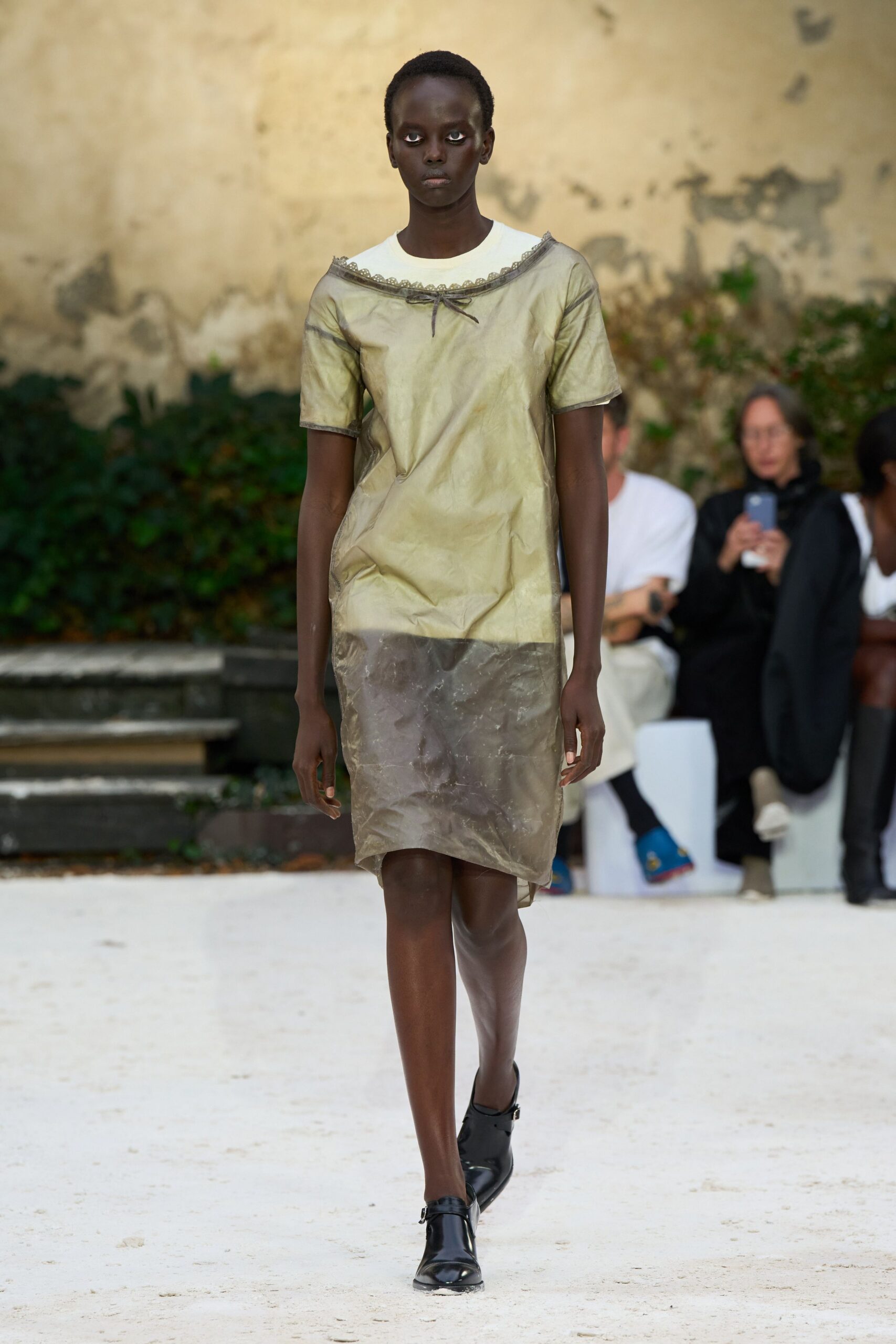 Crinkled metallic shift dress with a smoke-grey sheer hem and scalloped trim. White crewneck tee peeking cleanly above the off-shoulder neckline. Black Mary Jane flats with squared toes and a tiny buckle. Crossing a gravel courtyard before the museum opens after rain.