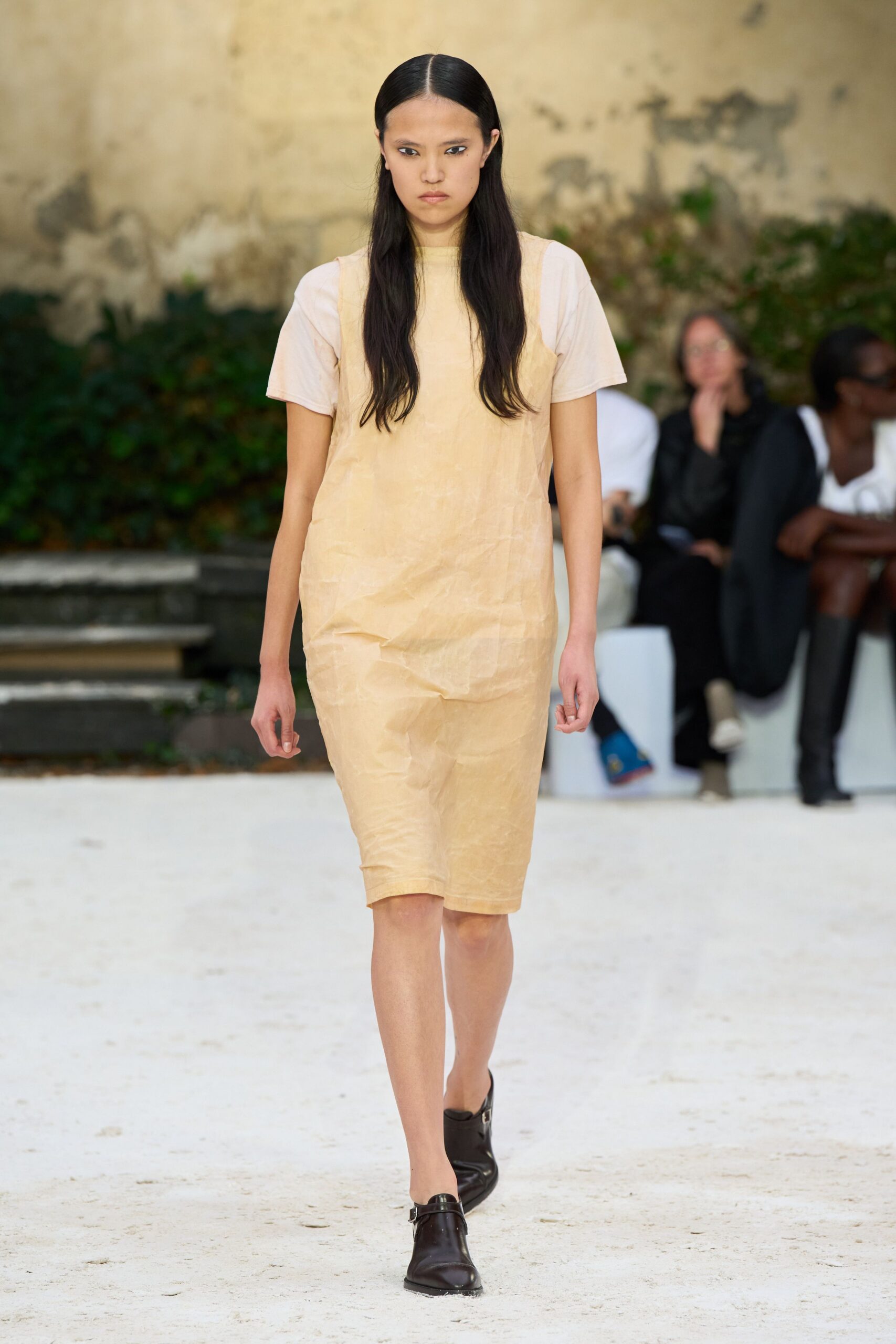 Pale apricot sheath dress in crinkled, papery fabric, hem stopping at the knee. Chalk short-sleeve tee layered underneath, neckline sitting close and flat. Black leather mules with squared toes and a slim strap buckle. Crossing a sunstruck cloister courtyard before the stone gives back the heat.