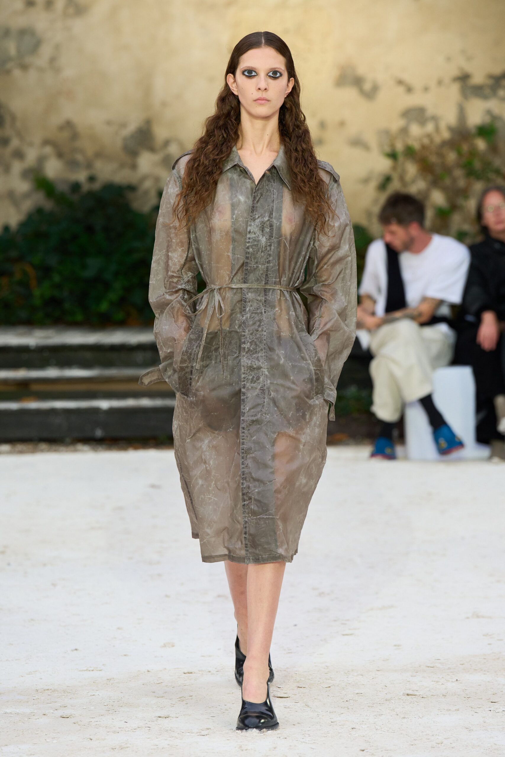 Smoky sheer coatdress in crinkled film, cinched with a narrow self-tie. Dark briefs visible beneath the translucent placket. Black pumps with a blunt squared toe. Crossing a courtyard just after the sprinkler shut off.