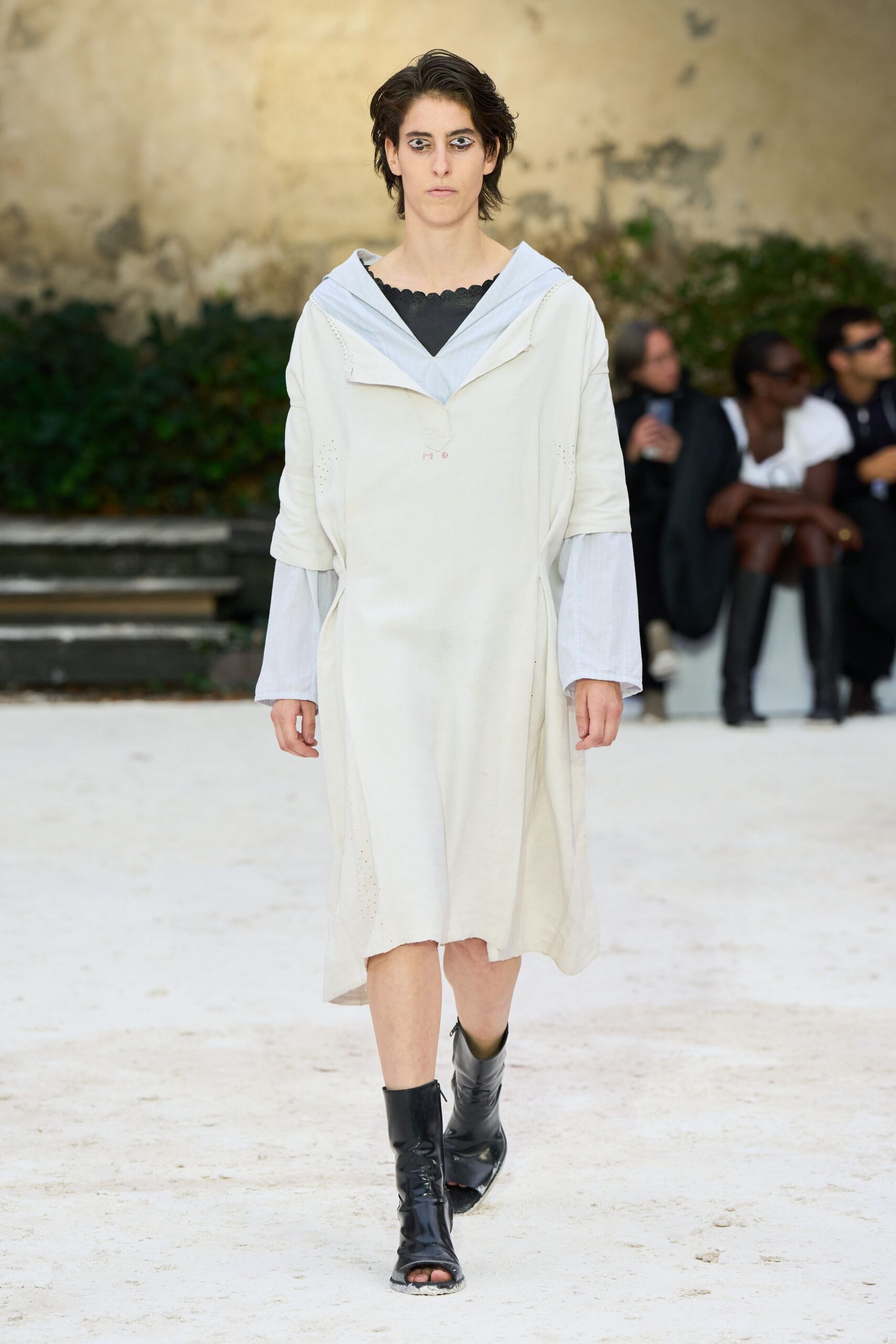 Chalk tunic dress with a sailor collar and tiny laddered seams. Pale blue gauze sleeves and hood peeking from beneath. Black scalloped neckline cutting a dark triangle at the chest. Black leather peep-toe boots, dust whitening the front edge. Crossing a monastery courtyard just after the stone floors were swept.
