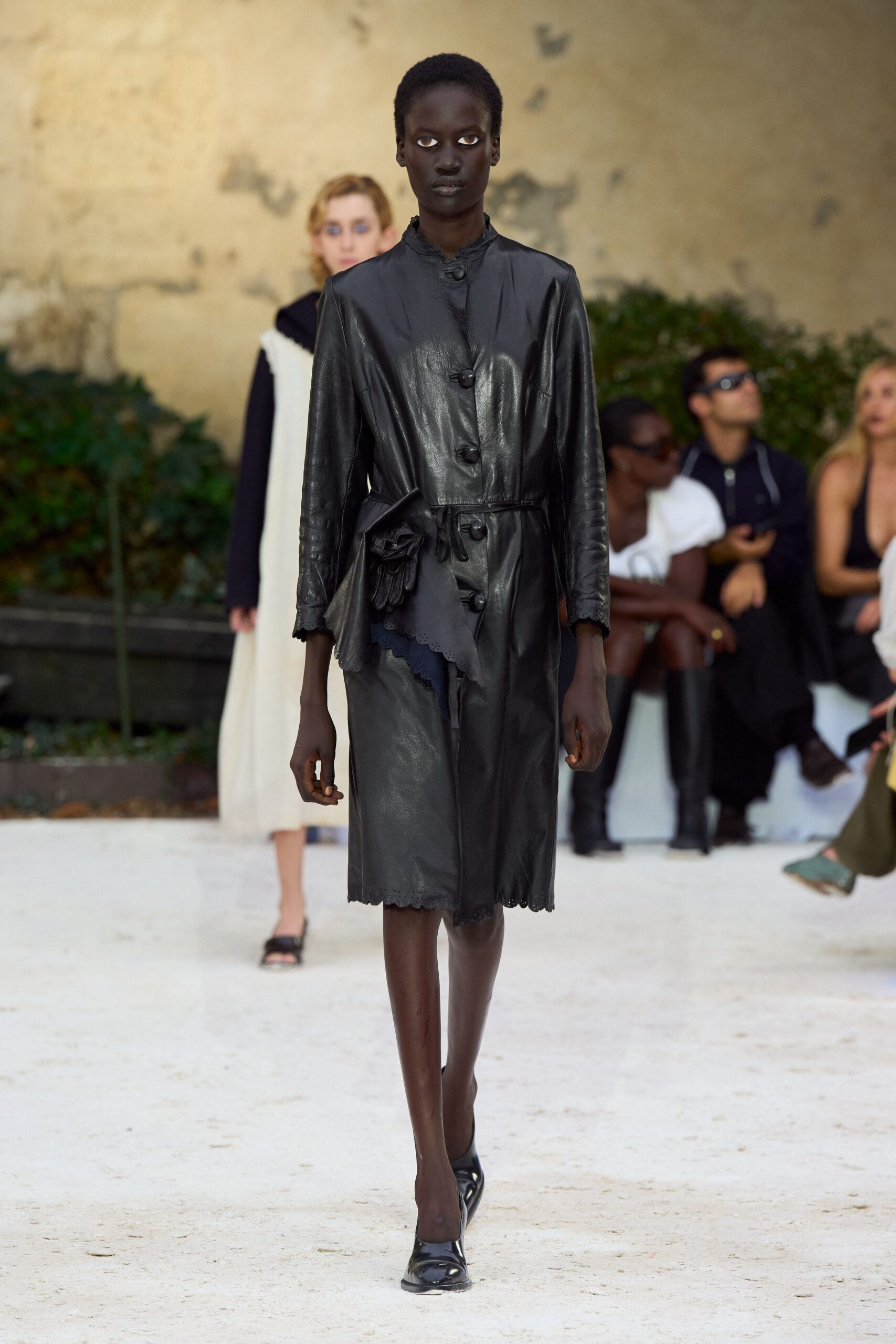 Black leather coat dress with scalloped eyelet hem and ruffled stand collar. One black glove tucked into the waist tie, fingers dangling. Black pumps with a softly squared toe. Crossing a gravel courtyard before the old villa doors are unlocked.