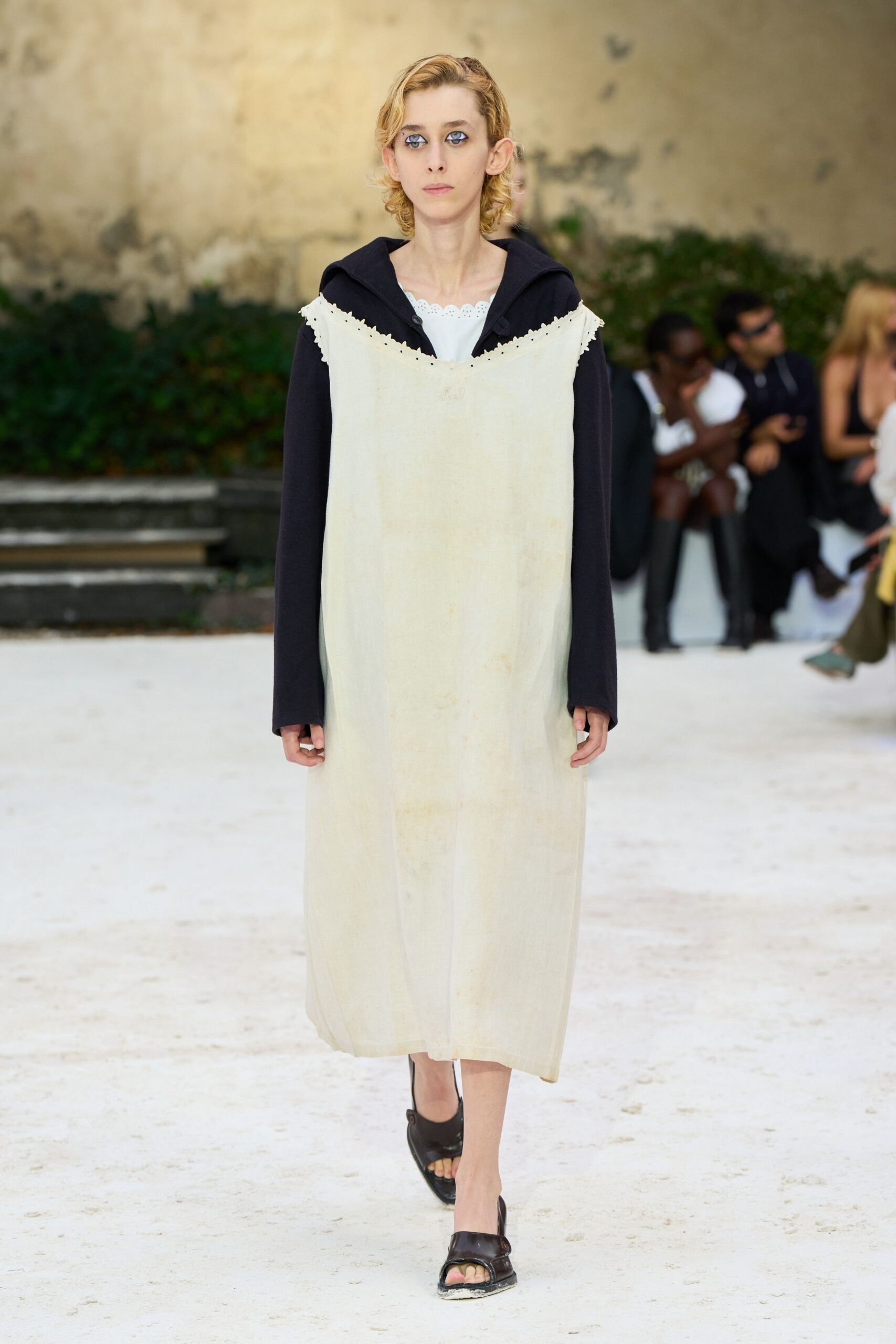 Long chalk dress with a scalloped eyelet bib laid over black sleeves. White underlayer peeking at the neckline in a neat scalloped strip. Black open-toe mules with thick uppers and dusty soles. Crossing a convent courtyard just after the whitewash dried.