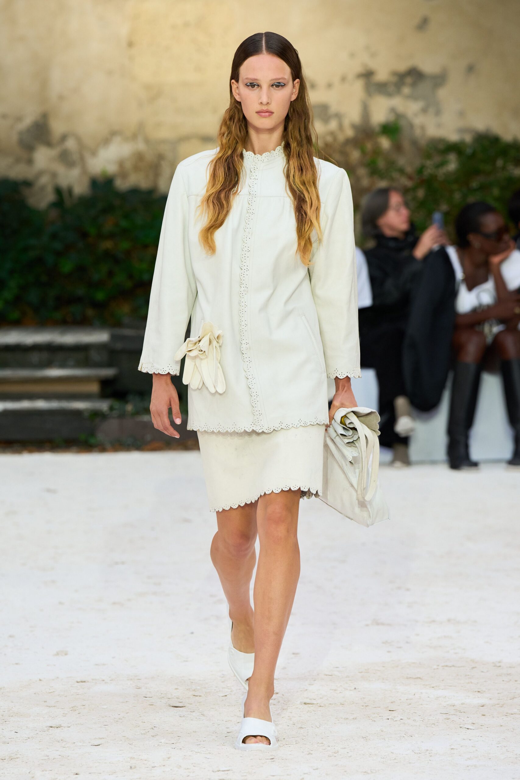Ivory leather tunic with scalloped eyelet edges and a glove rosette. Matching knee-length skirt, scalloped hem sitting just above the knee. Soft folded leather tote carried loose in one hand. White leather slide mules with a squared open toe. Crossing a gravel courtyard after the ceremony, bouquet already set down.