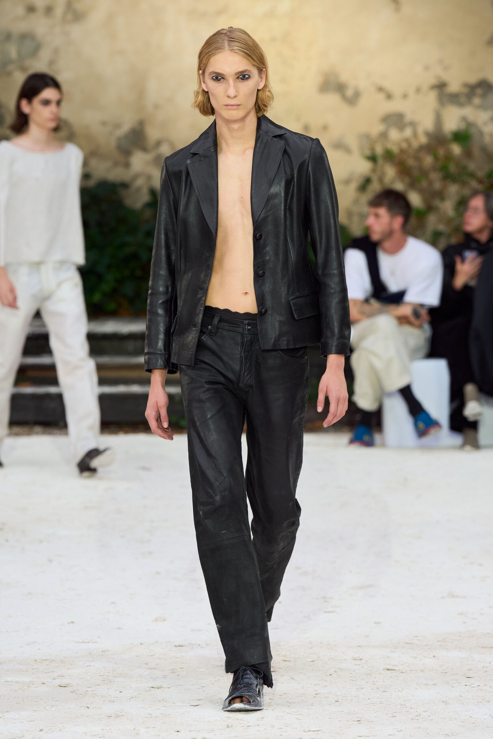 Black leather blazer worn bare, narrow lapels and matte horn buttons. Matching leather trousers, low rise with whiskering through the fly. Black split-toe slip-ons, silvered and scraped across the front. Walking through a crumbling courtyard before the heat has left the stone.