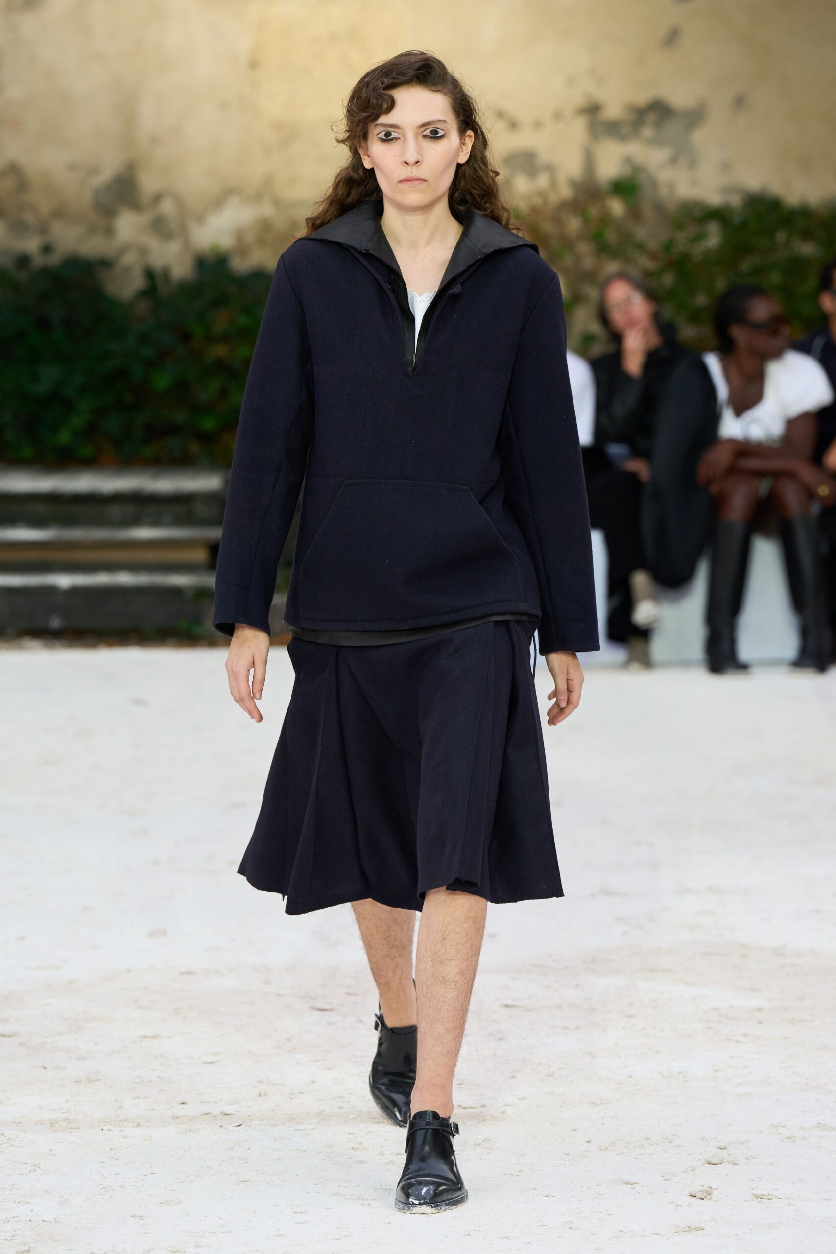 Ink wool anorak with a matte funnel collar and kangaroo pocket. White scalloped edge peeking from the half-zip opening. Black knee-length culottes cut with deep swinging folds. Black leather monk shoes, toes dusted chalky white. Crossing a monastery courtyard just after the hose water dries.