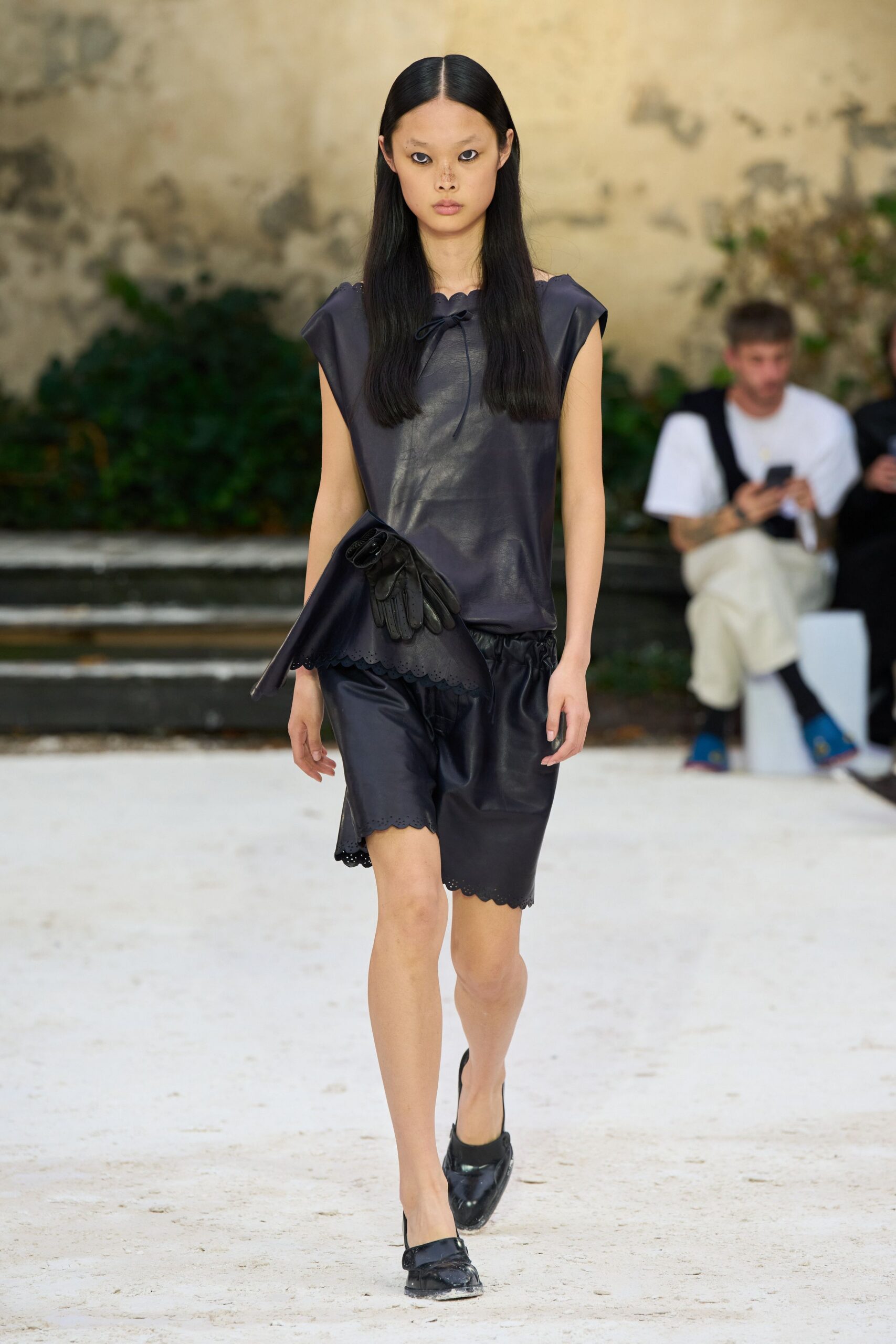 Ink leather shell top with scalloped neckline and a loose bow tie. Black leather driving gloves tucked under the arm. Matching leather knee shorts with broderie-scalloped hems and a gathered waist. Glossy black pointed mules, dusty at the tip. Crossing a gravel courtyard before lunch is called from inside.