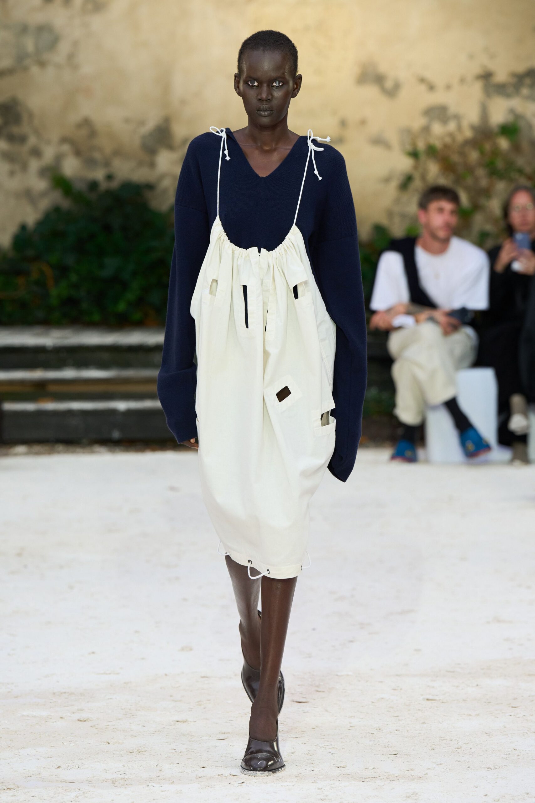 Navy knit sweater with a raw V neck and extra-long sleeves. Ivory cotton slip dress ruched on cord straps, patched slit pockets cut through. Dark square-toe flats, glossy and close to the foot. Walking across a courtyard before the laundry line is taken down.