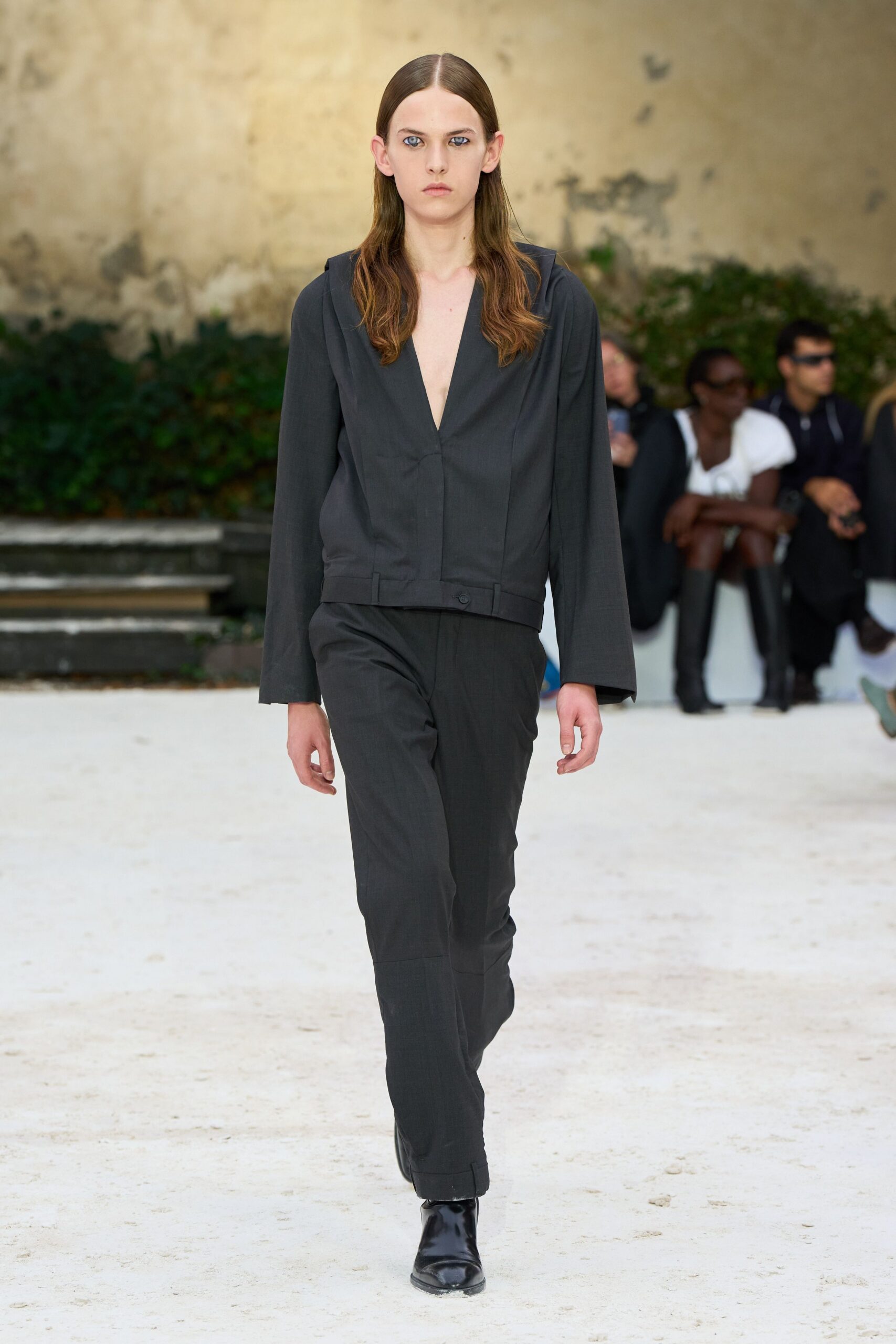 Charcoal suiting top with a plunging V and hidden front closure. Matching straight trousers, lightly pooled, with a broad cuffed hem. Black leather ankle boots with a blunt square toe. Crossing a gravel courtyard before the meeting turns into dinner.