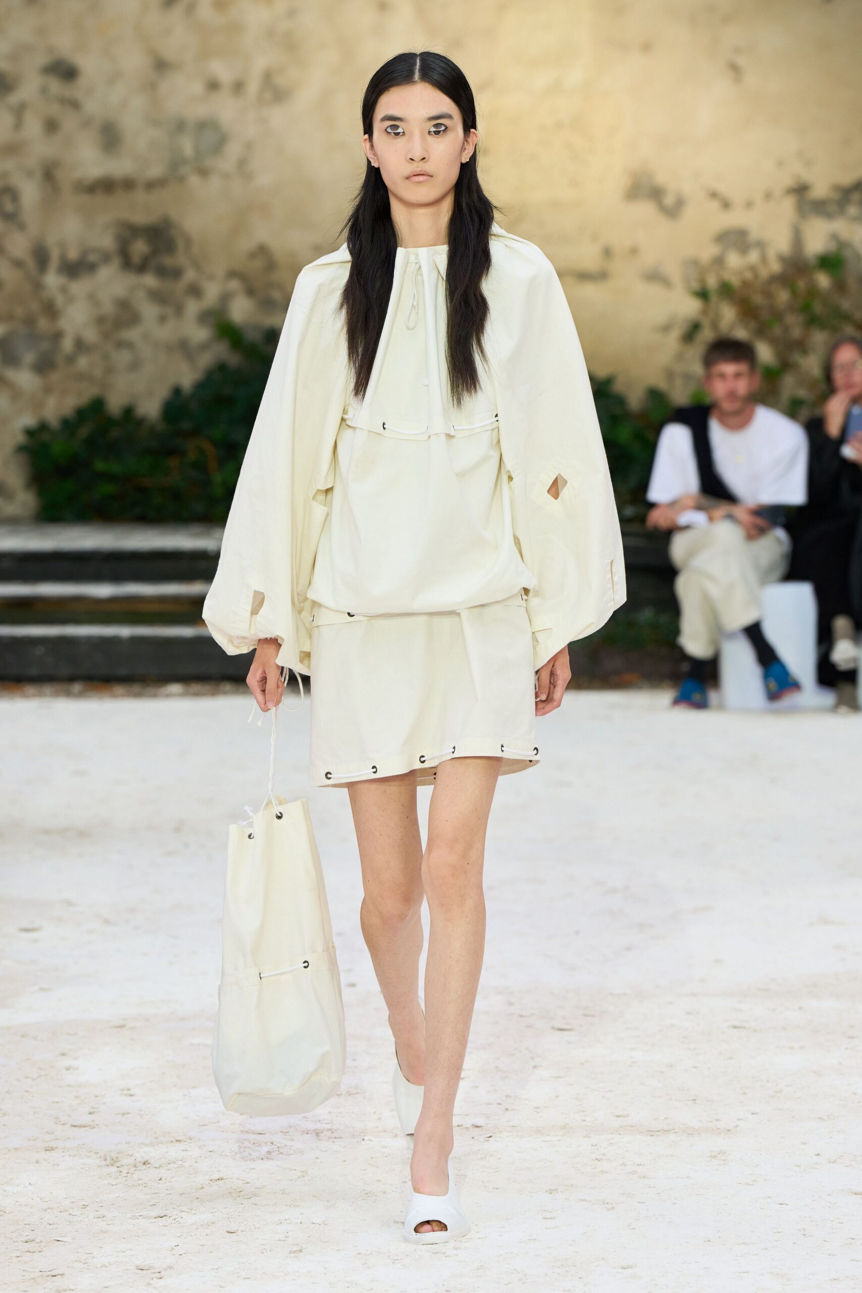 Cream cotton cape blouse with a gathered neckline and slashed sleeves. Matching drop-waist mini, hem threaded through black metal eyelets. Tall cream tote with the same corded eyelet seam. Milk-white peep-toe mules. Walking across a chalk courtyard before the heat lifts.