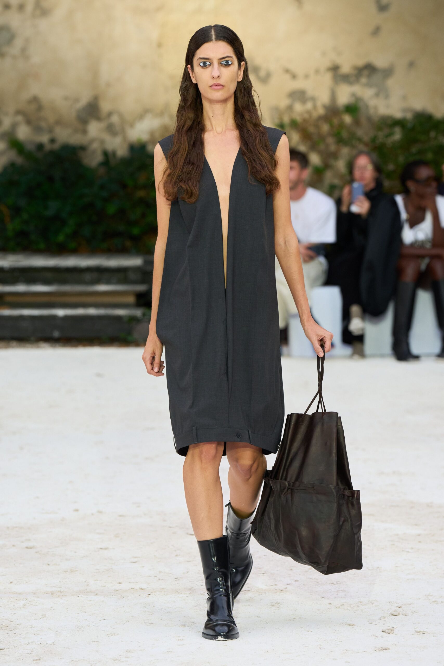 Charcoal sleeveless shift dress with a plunging split front and tabbed hem. Oversized black leather tote, side pockets slouching below the handles. Glossy black mid-calf boots with squared-off toes. Crossing a sunstruck cloister courtyard with somebody else's overnight things.