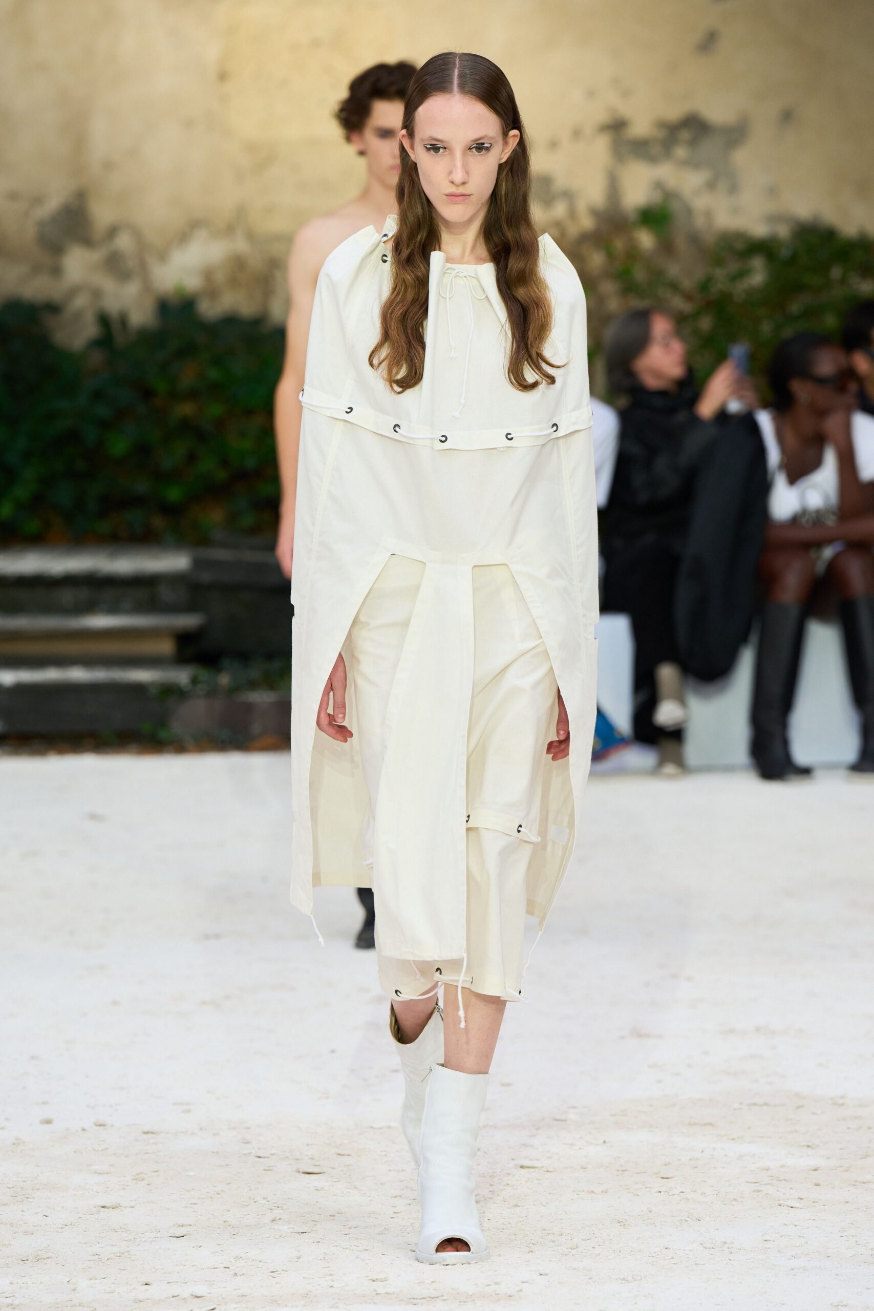 Milk-white tunic with metal grommets and drawcords skimming the chest. Matching cropped trousers with split apron panels and a dangling cord end. White peep-toe boots with a rubbery tubular shaft. Crossing a monastery courtyard just after the paving stones dried.