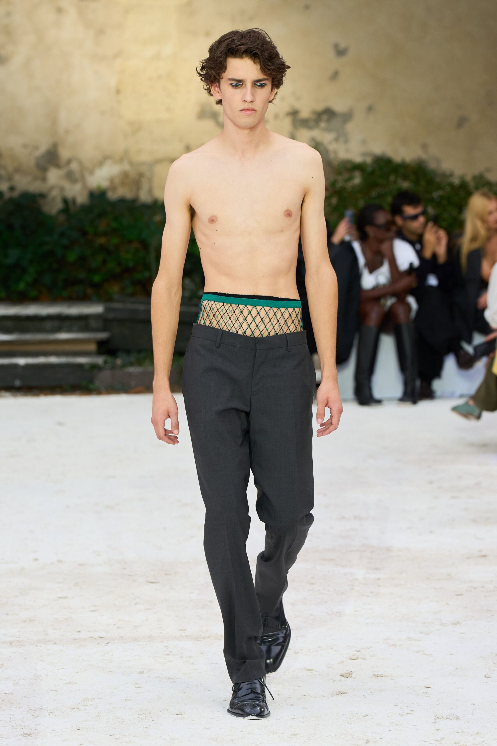 Sheer diamond-net brief with a deep green elastic top edge. Charcoal wool trousers riding low, waistband dropped beneath the mesh. Black derbies with a cloudy scuff across one toe. Crossing a gravel courtyard before anyone has opened the shutters.