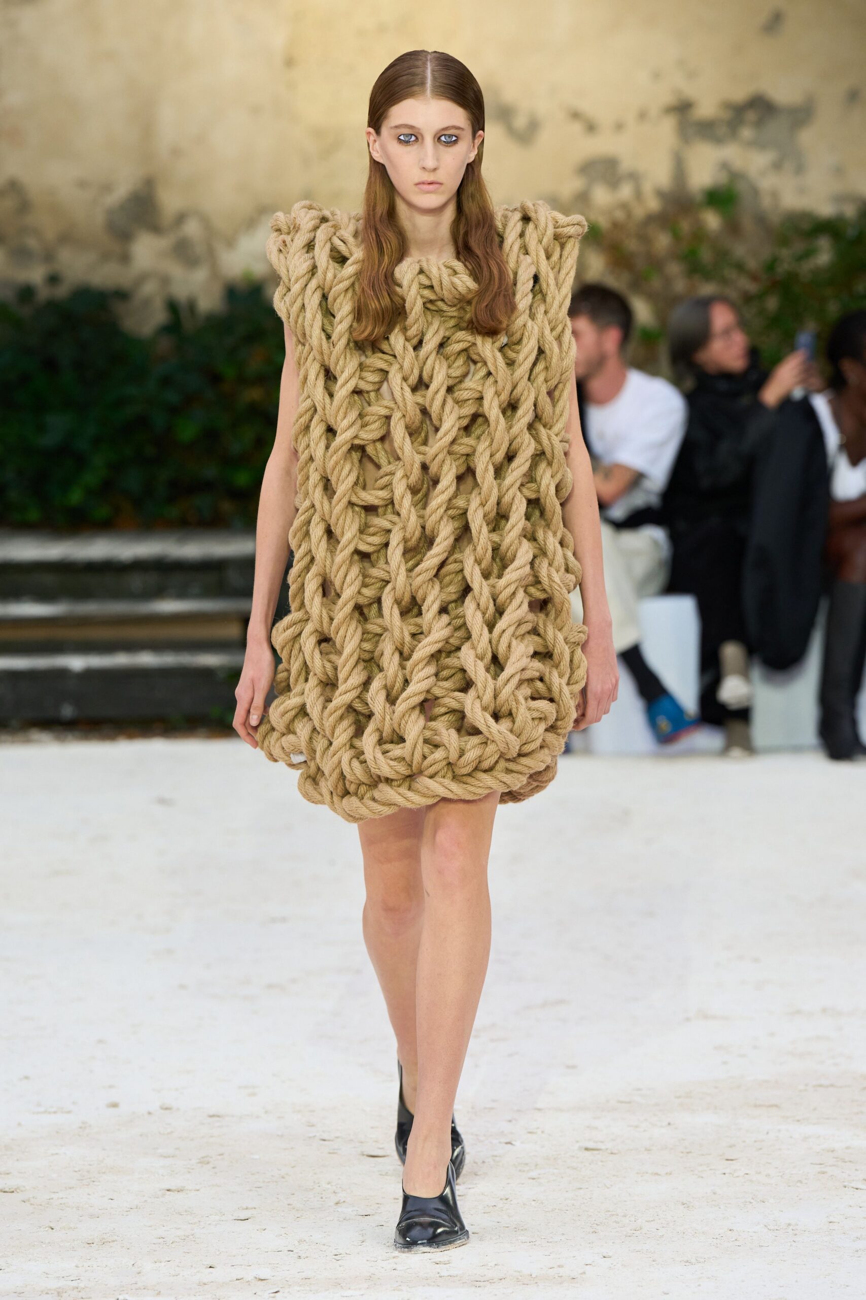 Sleeveless shift built from giant braided hemp rope, looped into a dense lattice. Black leather pumps with blunt square toes and scuffed tips. For crossing a museum courtyard after rain, still smelling the rope.