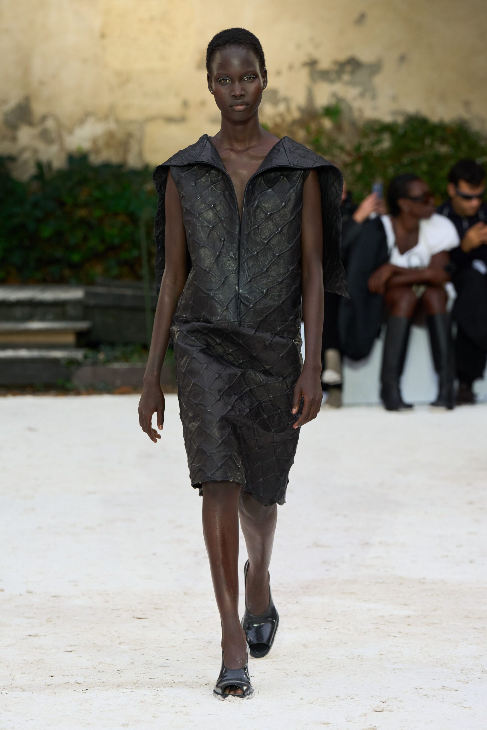 Black leather top pieced in oversized scale-like panels, zip splitting the plunging front. Matching knee skirt, the patchworked scales buckling slightly at one thigh. Black peep-toe heels with blunt squared vamps. Crossing a sunburnt courtyard after the fountain has run dry.