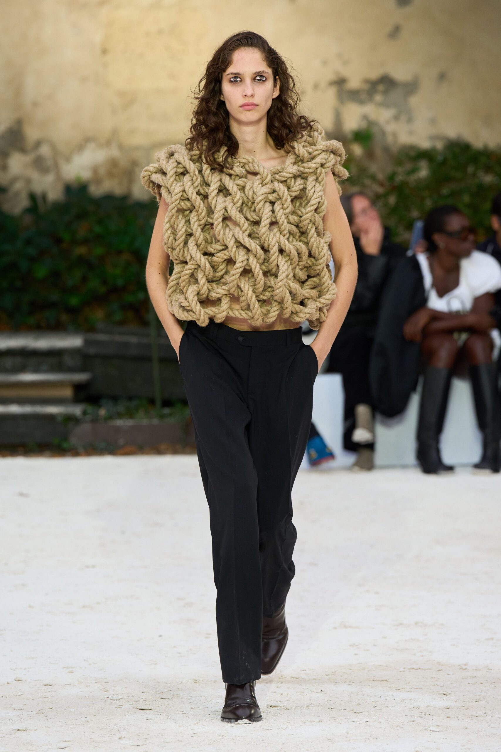 Openwork top built from giant natural rope braids, shoulder loops jutting outward. Black tailored trousers with a slanted closure and soft front crease. Dark leather ankle boots with squared toes peeking under the hem. Walking across a gravel courtyard after helping pull in the boats.
