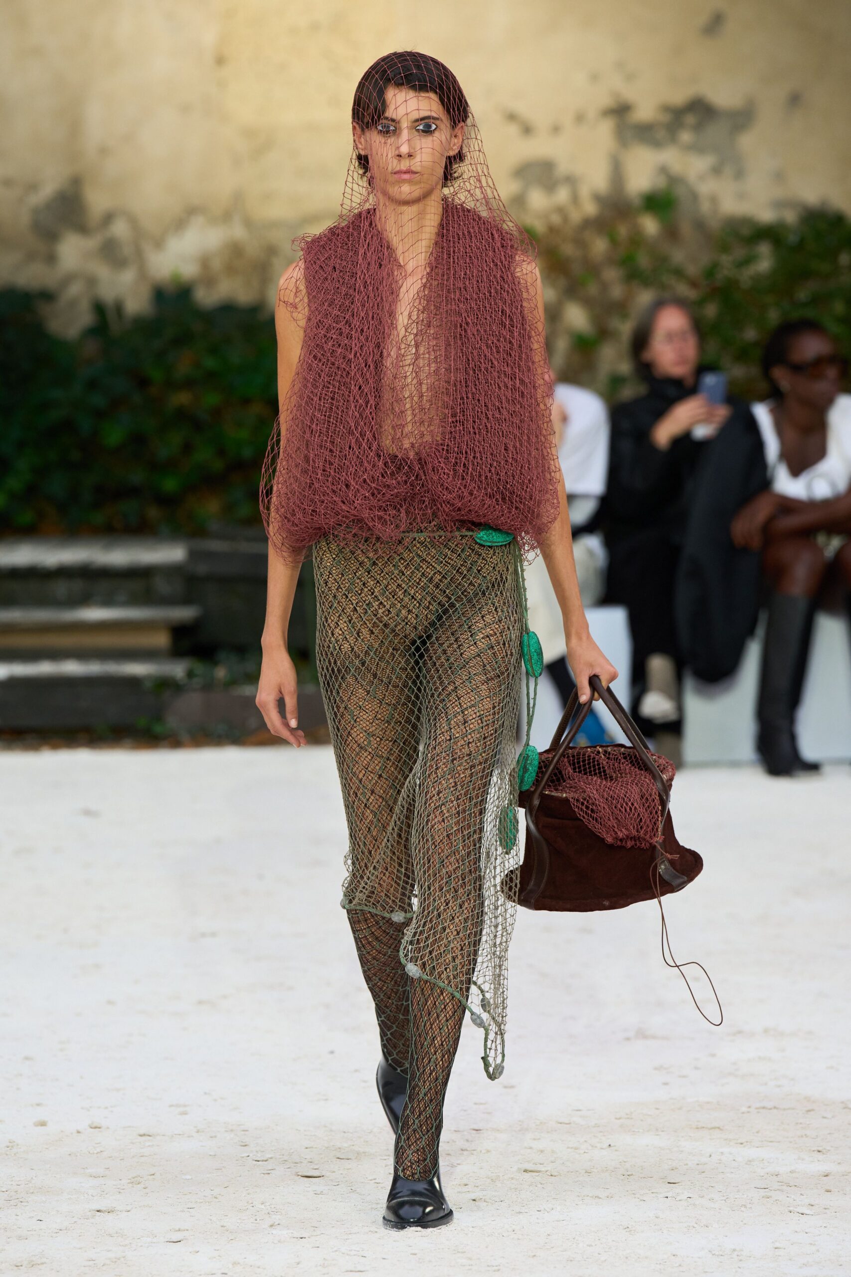 Burgundy open net draped from the head, pooling into a cowl front. Diamond-mesh trousers with green appliqué discs and a loose corded hem. Brown suede holdall snagged with a scrap of matching net. Black pointed boots, glossy at the toe. Crossing a dried fountain with someone's fishing nets still on you.