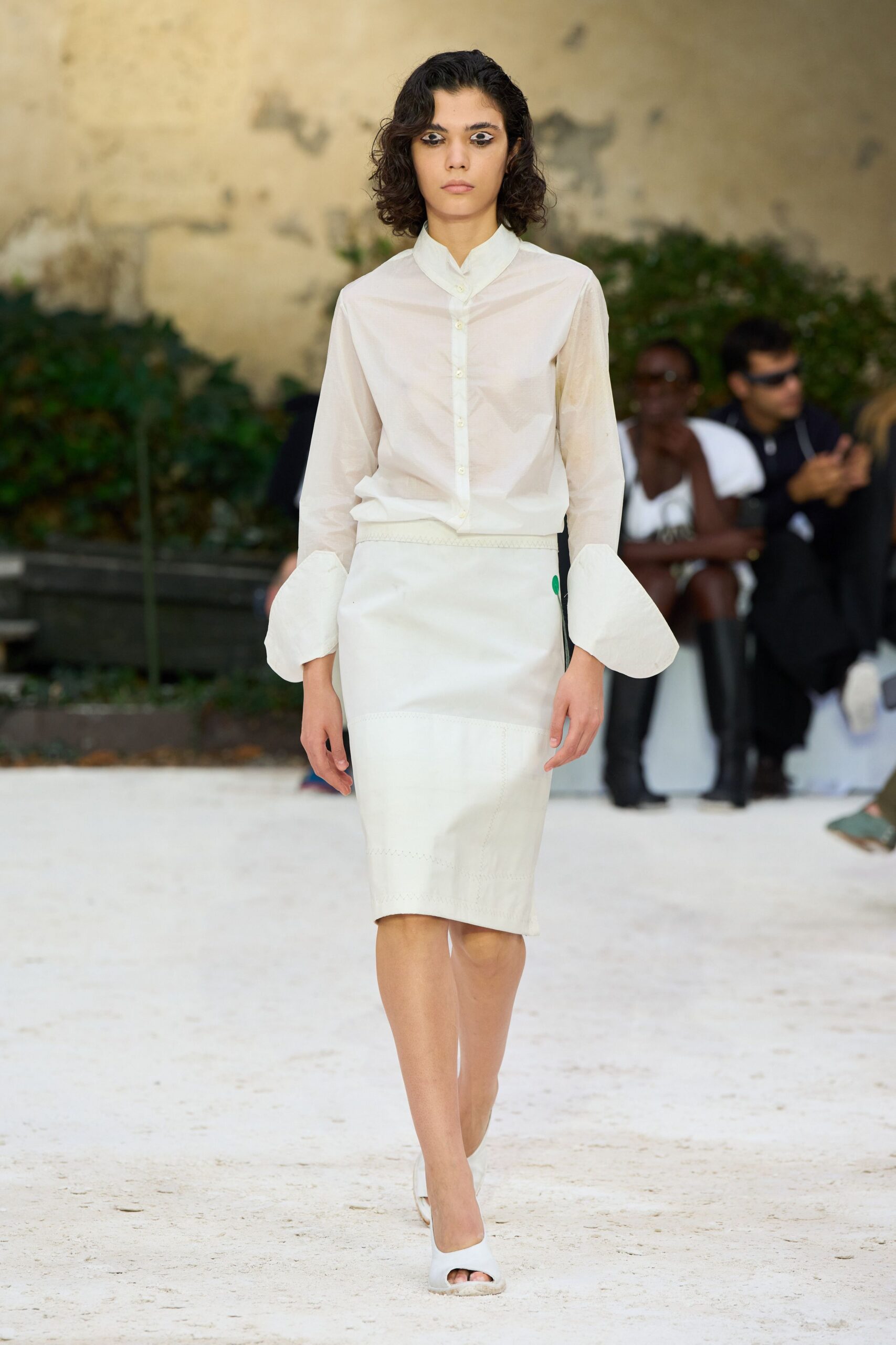 Sheer ivory blouse with a folded stand collar and ballooned cuffs. Milk-white pencil skirt seamed in broad panels, one green tab at the hip. White peep-toe mules with thick, softly rounded uppers. Crossing a convent courtyard just after the stones give back the heat.