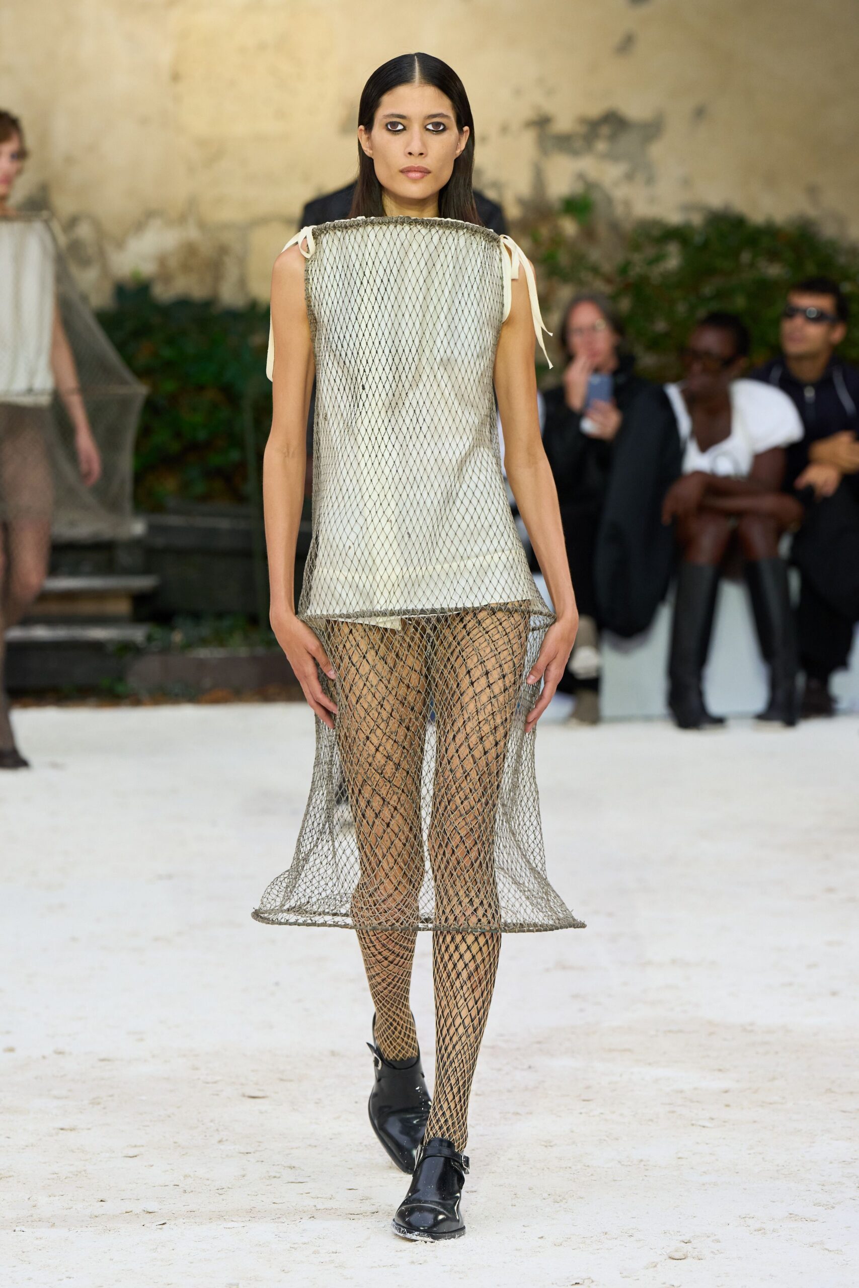 Rigid silver mesh tunic over a chalk slip, tied with narrow shoulder ribbons. Diamond-net tights turning denser around the calves. Black patent monk-strap flats with squared, slightly upturned toes. Crossing a sculpture garden before the rain starts.