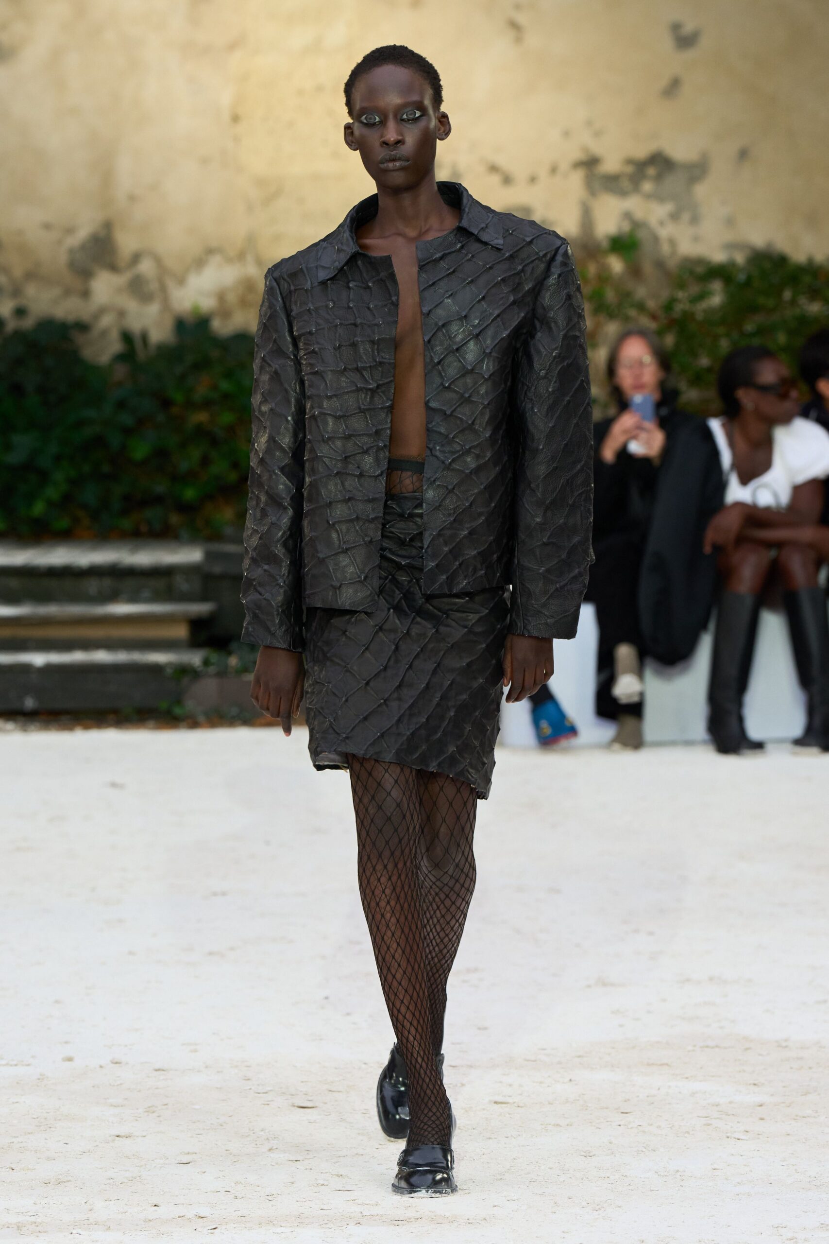Black jacket built from overlapping scale-like panels, plunging open at center. Matching black mini, wrapped askew in the same scaled construction. Wide diamond fishnet tights. Black loafers with a clear lucite sole edge. Crossing a gravel courtyard before rain, windows already dark at four.
