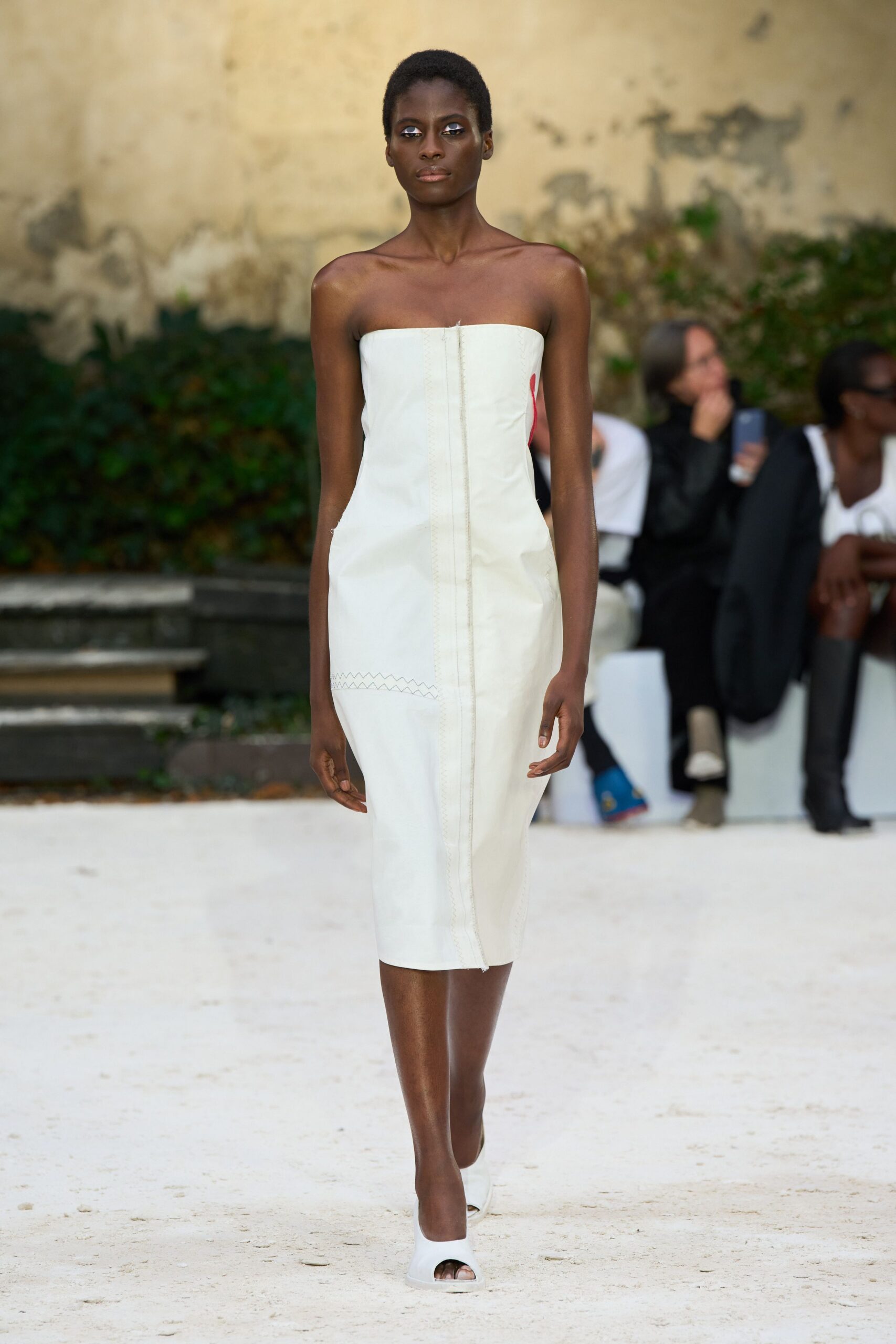 Strapless ivory column dress with exposed center seam and zigzag topstitching near the hip. White peep-toe mules with thick sculpted platforms. Crossing a chalk courtyard before the wedding guests have found their seats.