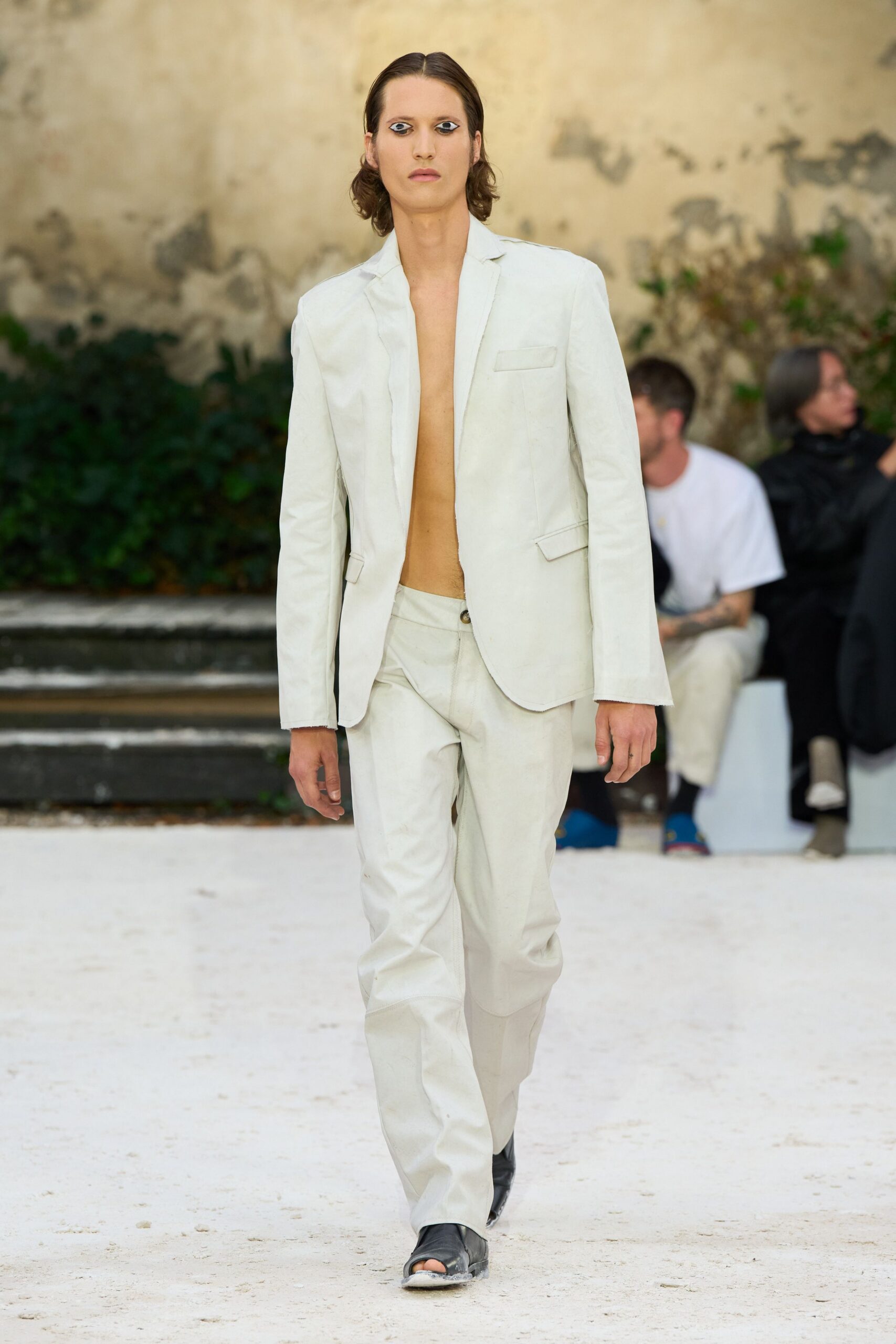 Chalk single-breasted blazer worn bare, with slightly raw cuff edges. Matching low-slung trousers with a dark horn button at the waistband. Black leather boots split open at the toe. Crossing a sunblown courtyard before the dust settles again.