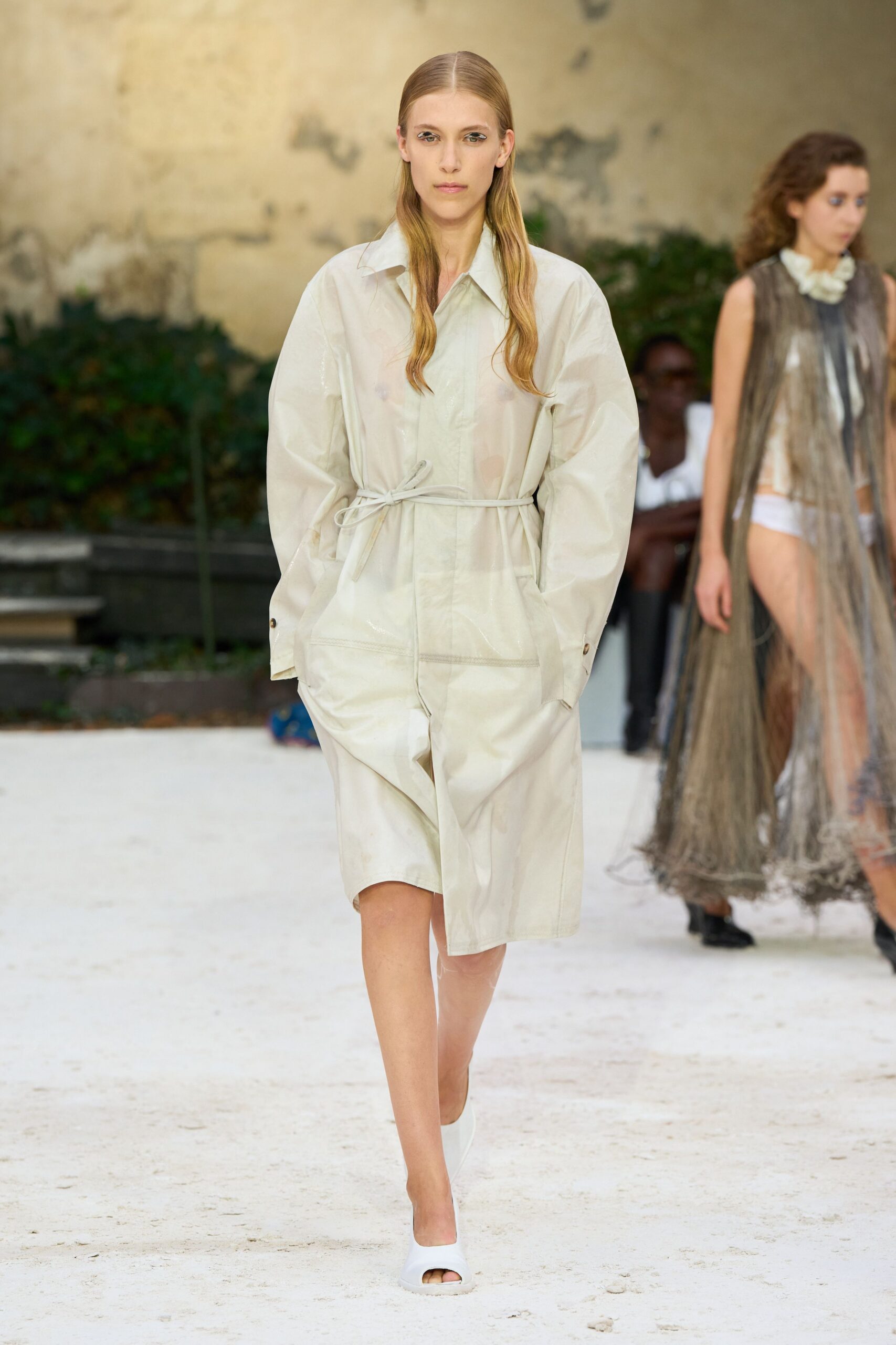 Translucent ivory shirt with a broad collar and balloon sleeves. Matching knee-length shorts in crisp coated cotton, softly creased. Thin self-tie cord knotted low at the waist. White peep-toe mules with a rounded rubbery vamp. Walking across a sunblown courtyard after swimming, still not fully dry.