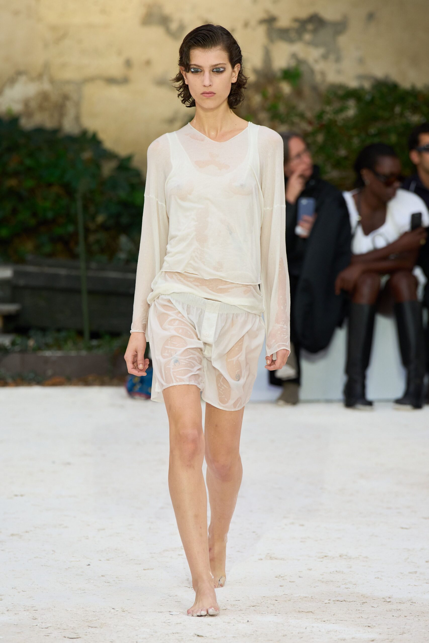 Gauzy ivory long-sleeve top, damp and clinging with a rolled hem. Pale scoop-neck tank ghosting through underneath. Sheer ivory shorts with an elastic waist and cloudy wet patches. Clear heels nearly disappearing against the sand. Walking back from the shore before your shirt has dried.