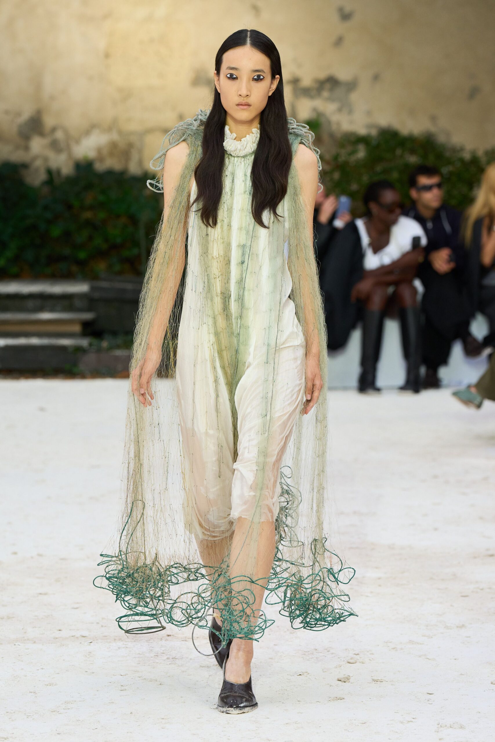 Ivory sleeveless dress with a ruffled stand collar and watery green streaks. Seafoam cord fringe knotted at the shoulders, dragging into loose coils. Black pumps dusted chalk-white at the toe. Crossing a dried fountain after the gardeners have rinsed the stone.