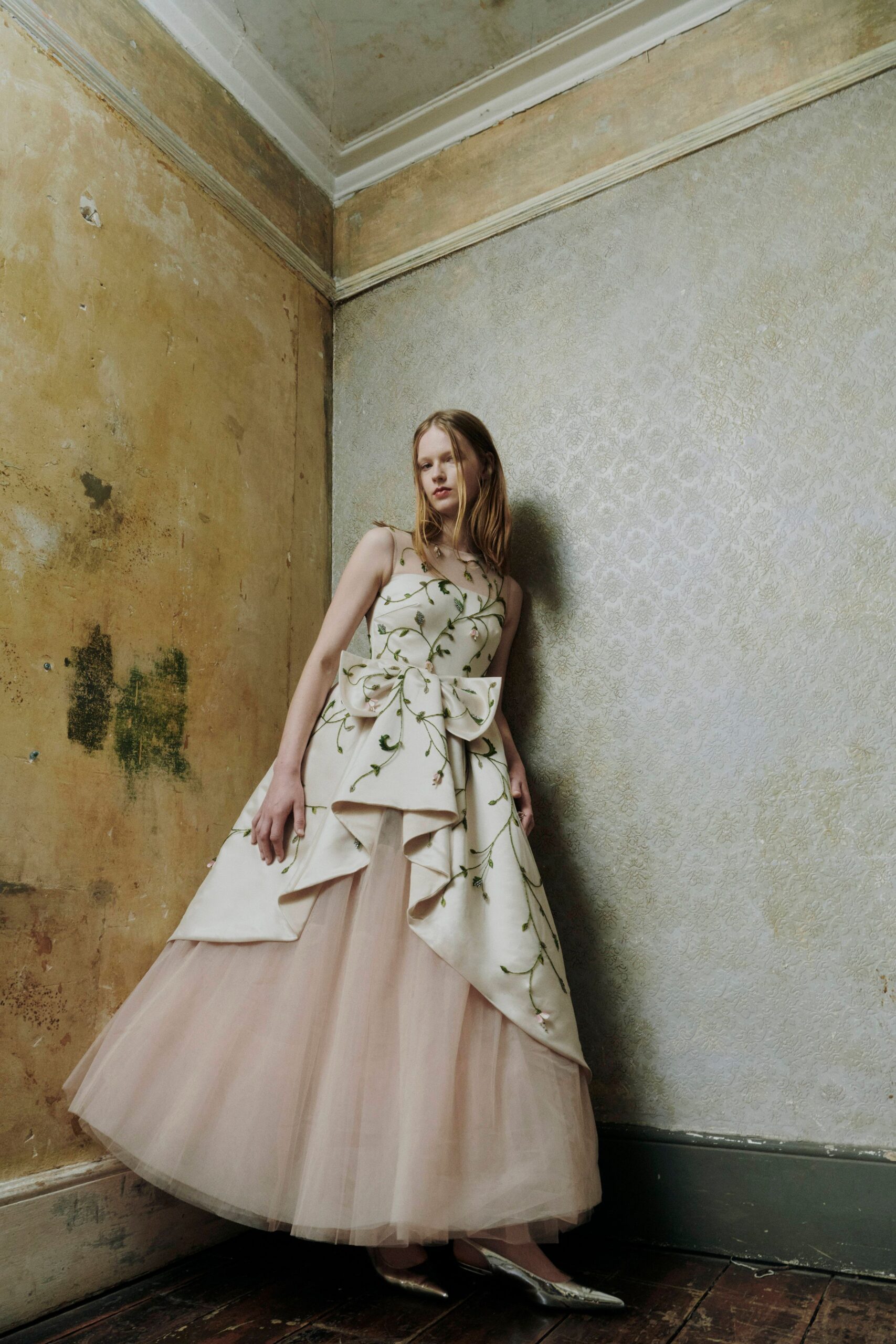 Ivory satin bodice and overskirt traced with green vine embroidery. Huge waist bow, one panel folding into a sharp draped point. Blush tulle ball skirt clouding out beneath the satin layers. Silver pointed pumps. In a faded manor drawing room after the flowers have started dropping petals.