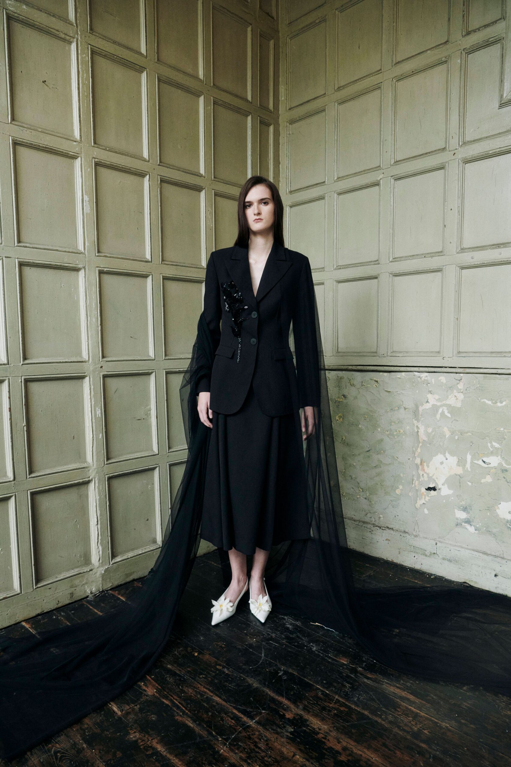 Black tailored blazer with peaked lapels and a sequined bloom climbing one side. Sheer black tulle train falling from the sleeves to the floorboards. Calf-length black skirt with a soft, weighty drape. White pointed pumps topped with crumpled petal rosettes. After the ceremony, alone in a paneled room that still smells of dust.