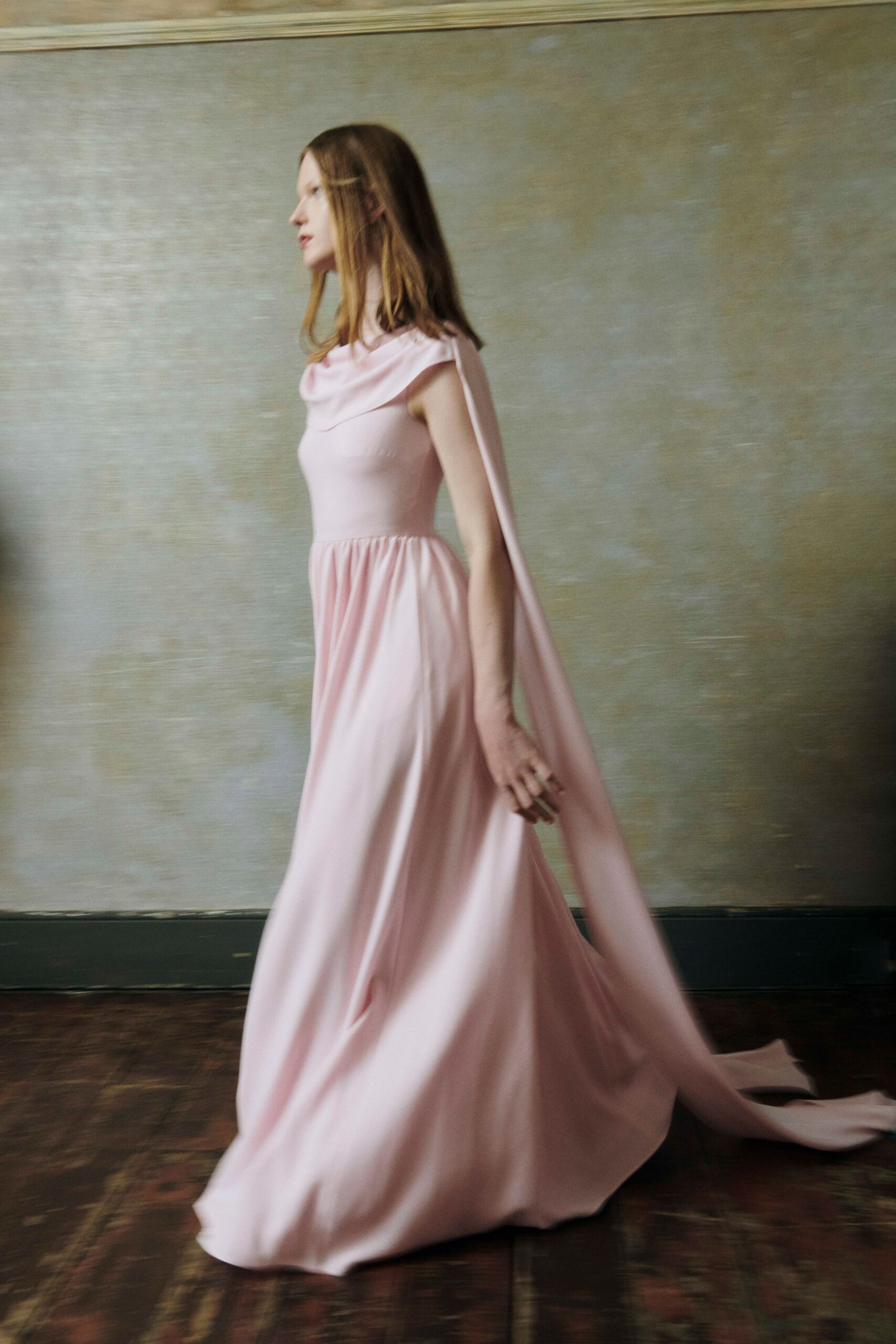Blush floor-length gown with a draped cowl neck and one cape panel trailing from the shoulder. Chunky gold ring catching at the swinging hand. Crossing a parquet room just after the vows, windows still open to the garden.