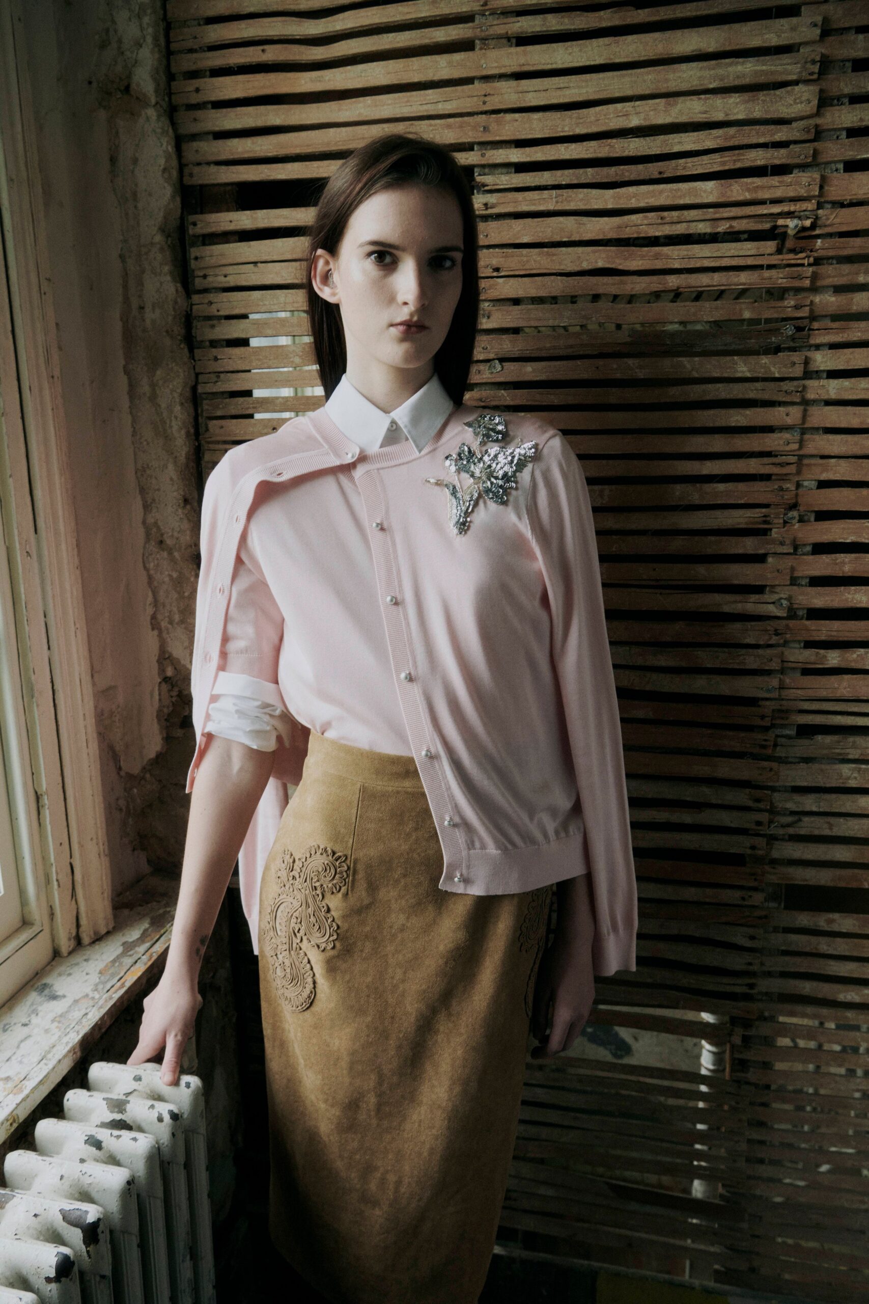 Blush fine-knit cardigan worn cape-style, pearl buttons and a silver beaded flower. White cotton shirt with a sharp collar and sleeves pushed to the forearm. Moss-brown suede pencil skirt with appliqued paisleys climbing one hip. By the cold window before a house viewing, radiator knocking under the sill.