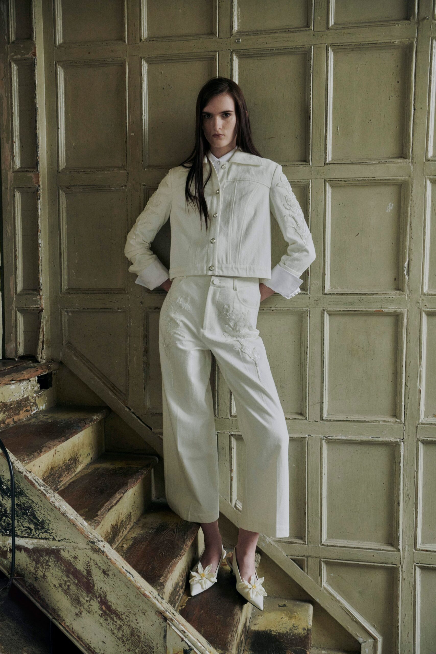 Cropped ivory jacket with pearl snaps and raised floral appliqué on the sleeves. High-waisted ivory trousers, pressed wide legs marked with scattered floral appliqué. Pointed satin mules topped with oversized petal rosettes. On the stair landing before a registry-office wedding in an old house.