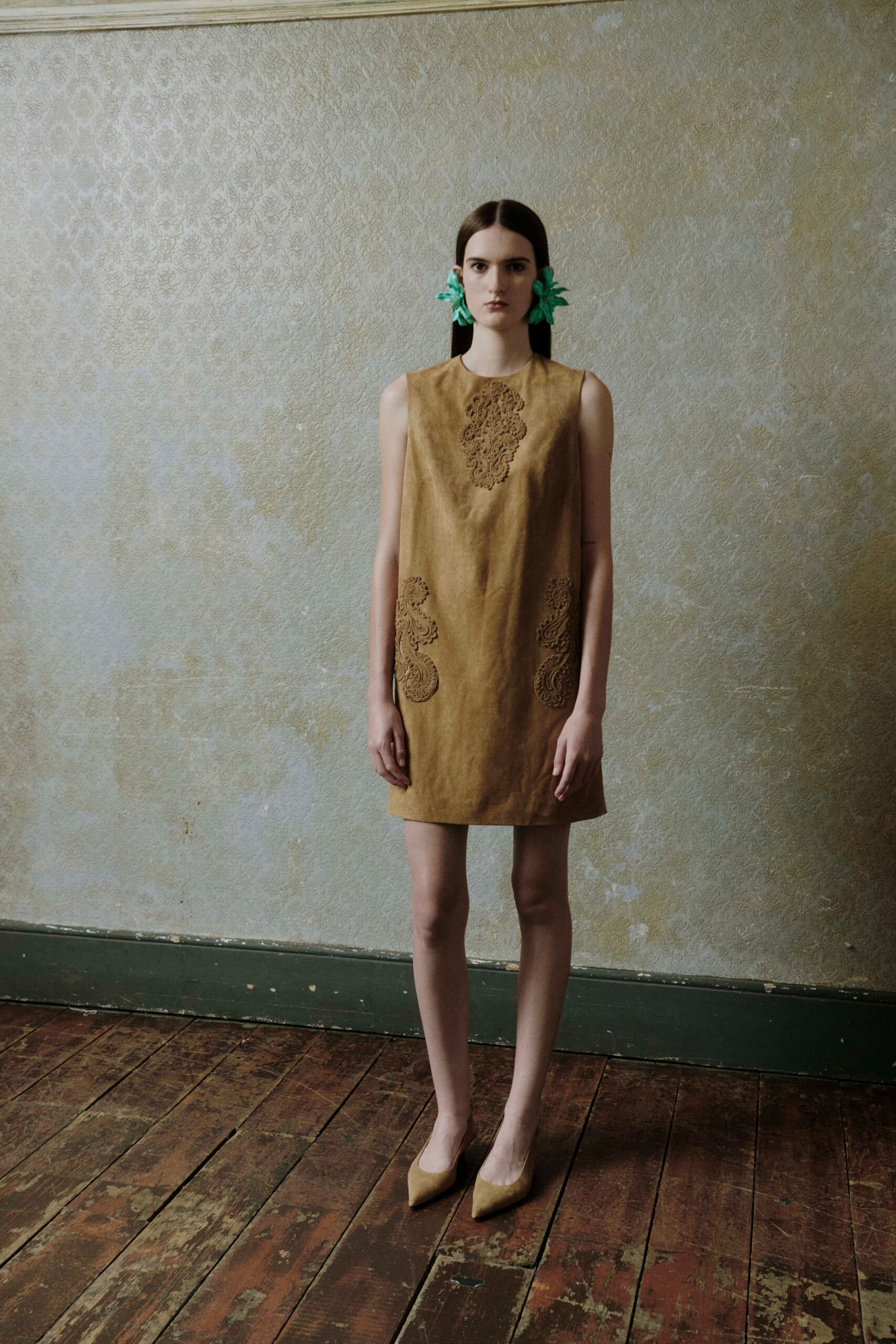 Mint green fabric flowers clipped low over both ears. Ochre suede shift dress with laser-cut scrollwork at chest and hips. Matching suede pumps with narrow pointed toes. Afternoon in a faded drawing room before the wedding guests arrive.