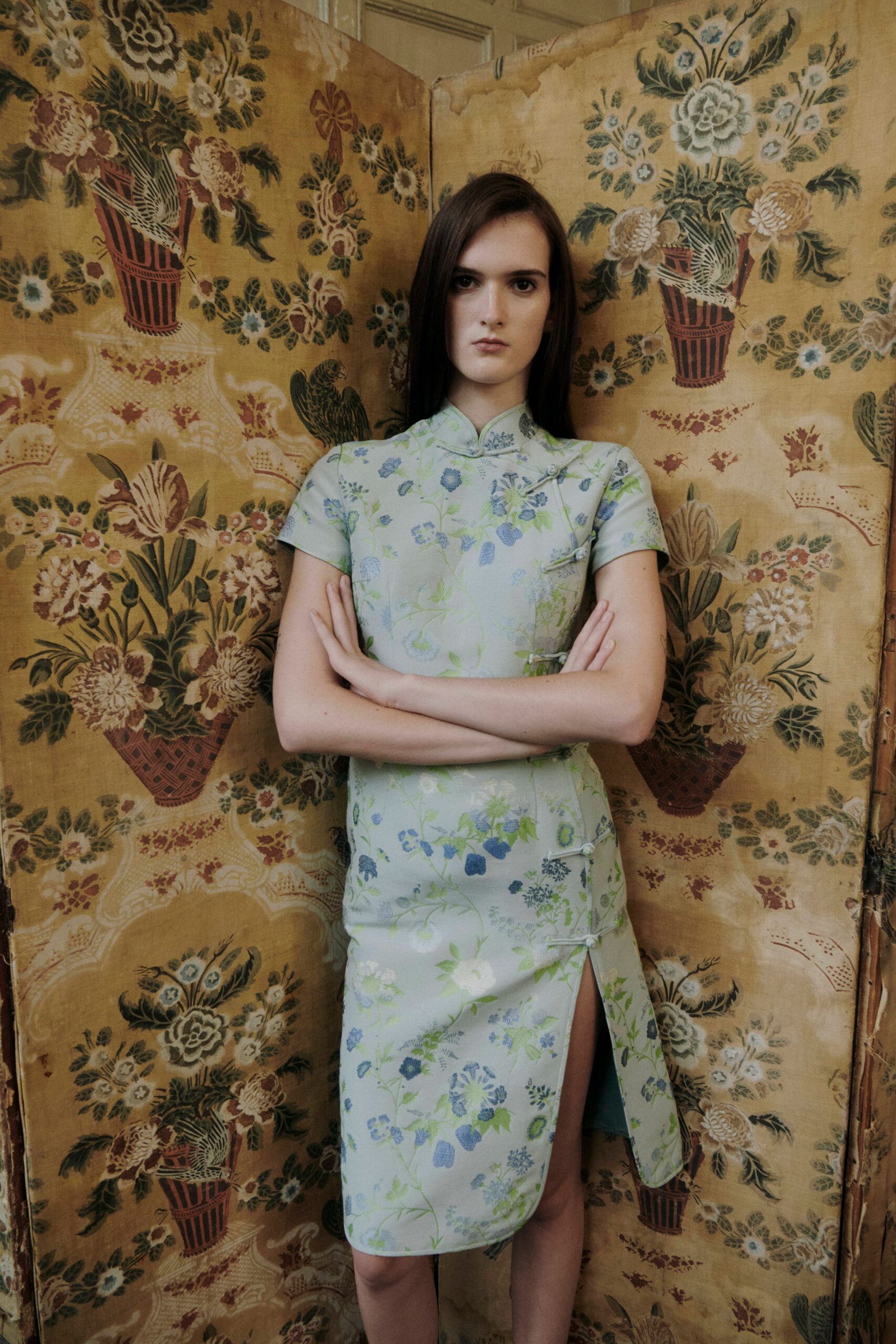 Pale celadon brocade cheongsam, blue floral sprigs and frog closures skimming sideways. High front slit opening from the lowest knot. Afternoon in a rented house, standing between folding screens and old wallpaper.