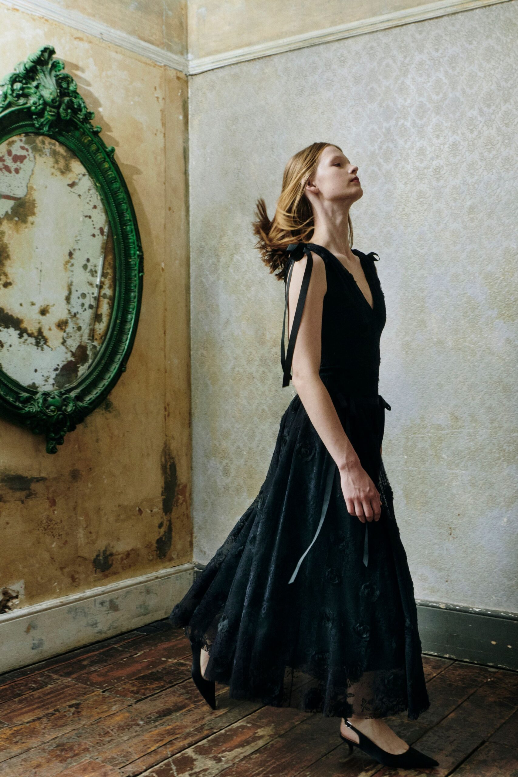 Black sleeveless dress with grosgrain shoulder ties and a sheer rosette-overlay skirt. Black pointed slingbacks with a small silver buckle at the heel. Crossing a drafty manor room before anyone has lit the fire.