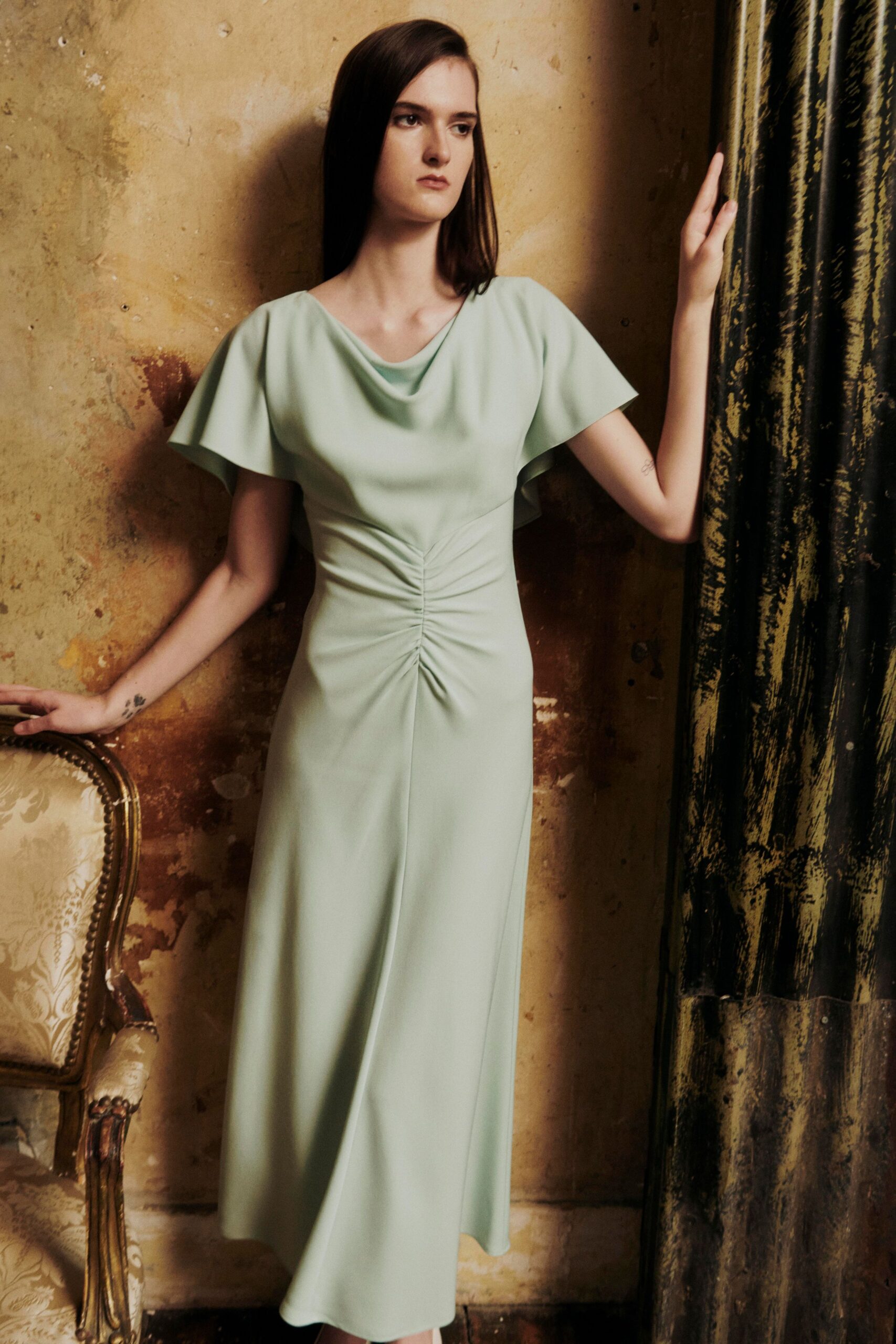 Pale celadon crepe dress with a draped cowl neck and ruched center seam. Soft flutter sleeves falling in shallow arcs from the shoulder. In a faded hotel parlour before someone opens the curtains.