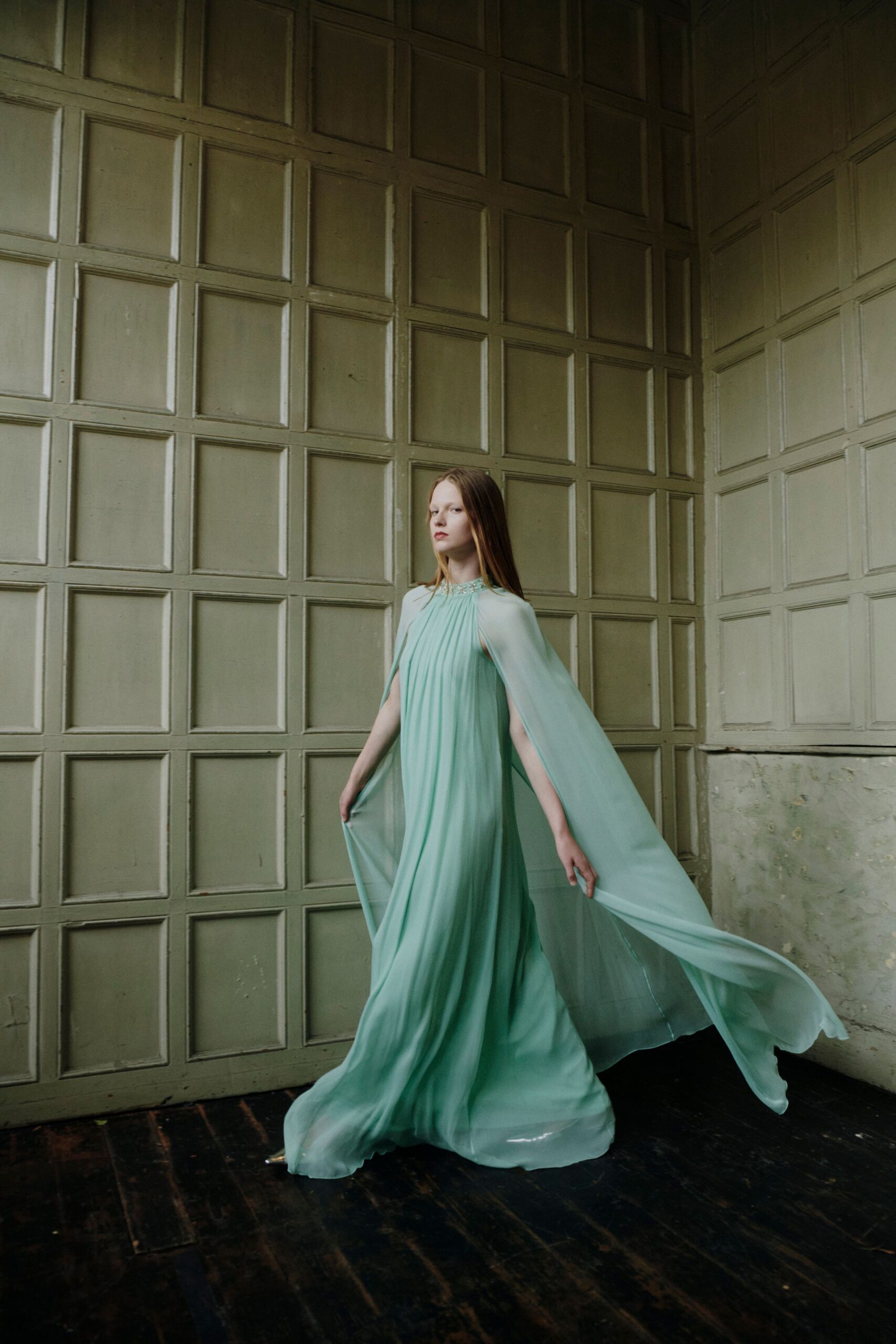 Seafoam chiffon gown, finely pleated from a jeweled neckline into cape sleeves. Silver pointed heels flashing under the pooling hem. Crossing a drafty manor hall before dinner is called.