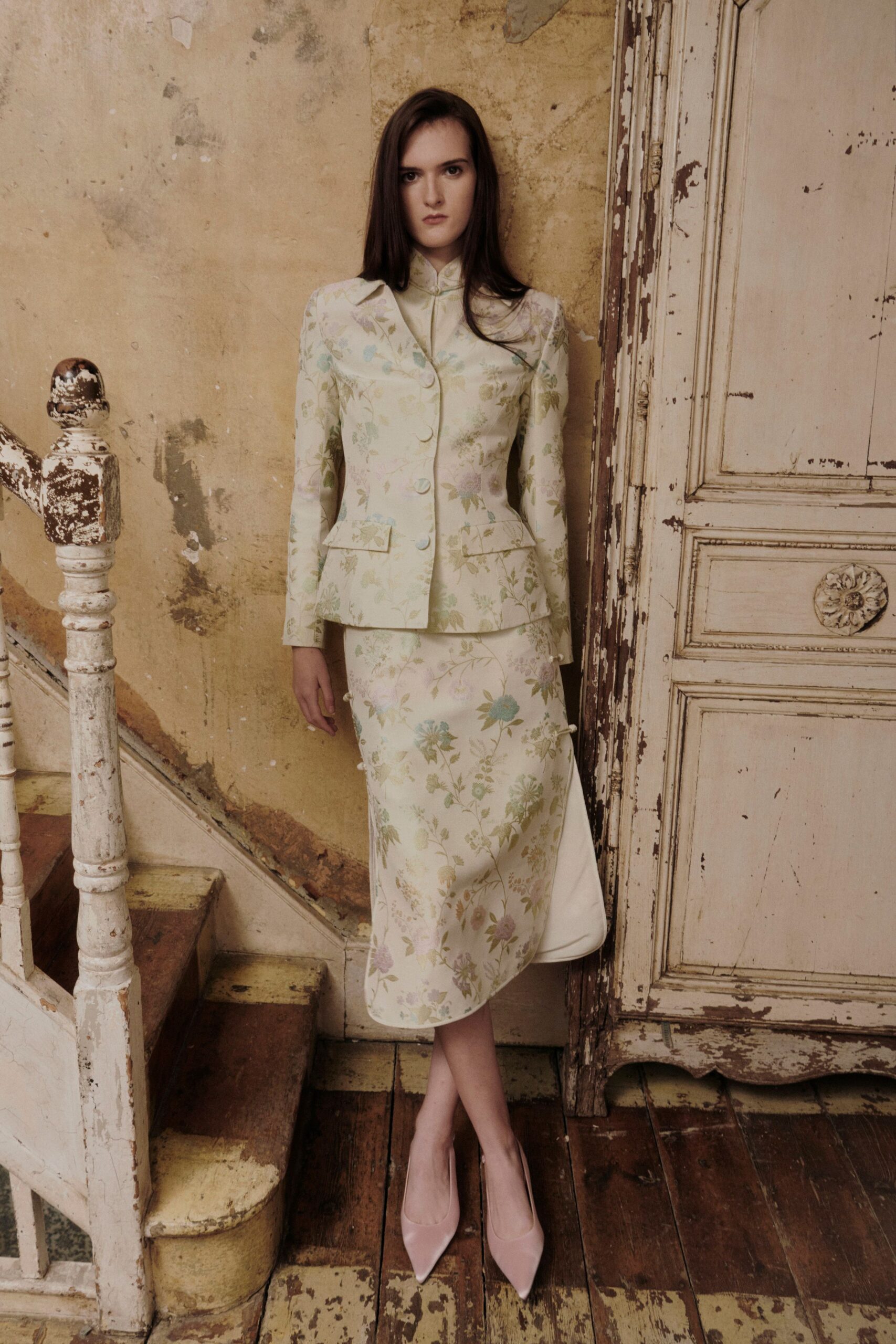 Cream satin blouse with a frog fastening at the throat. Fitted ivory jacket in washed floral brocade, flap pockets kicked outward. Matching midi skirt, side slit caught with tiny frog closures. Powder-pink pumps with sharp vamps. In a peeling stair hall after the estate agent has left.