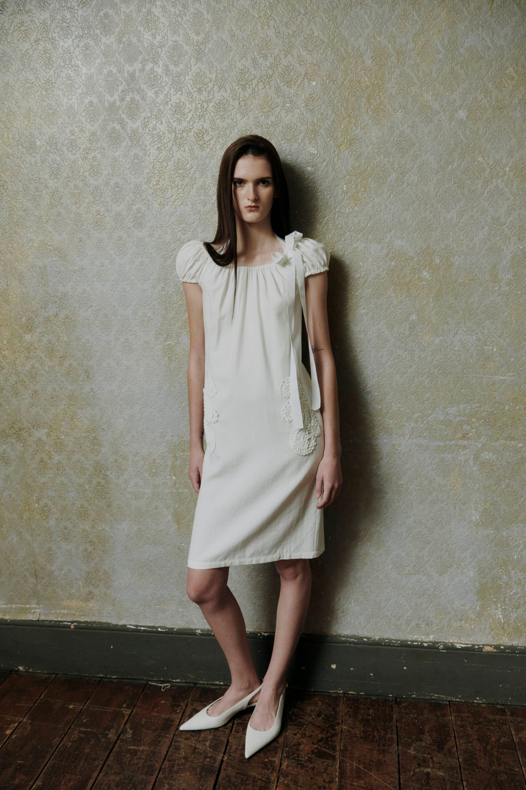 Ivory shift dress with puff sleeves, ribbon bow and trailing tie at one shoulder. White pointed pumps with a low-cut vamp. In a faded boarding-house hallway before somebody opens the parlor door.