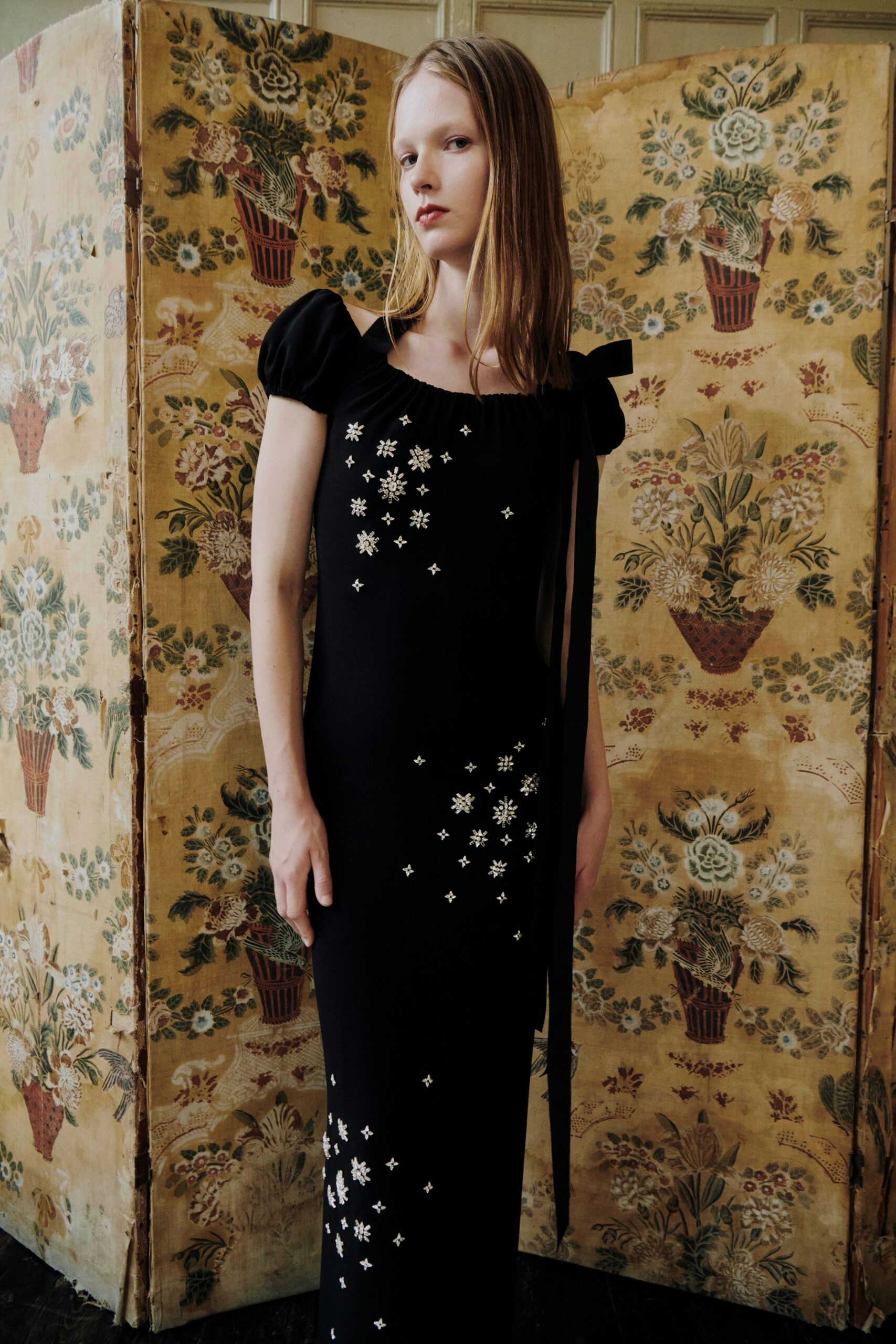 Black column dress with puff sleeves, crystal snowflakes scattered to the hem. Long black grosgrain tie falling from one shoulder bow. In a faded drawing room while the guests are still downstairs.