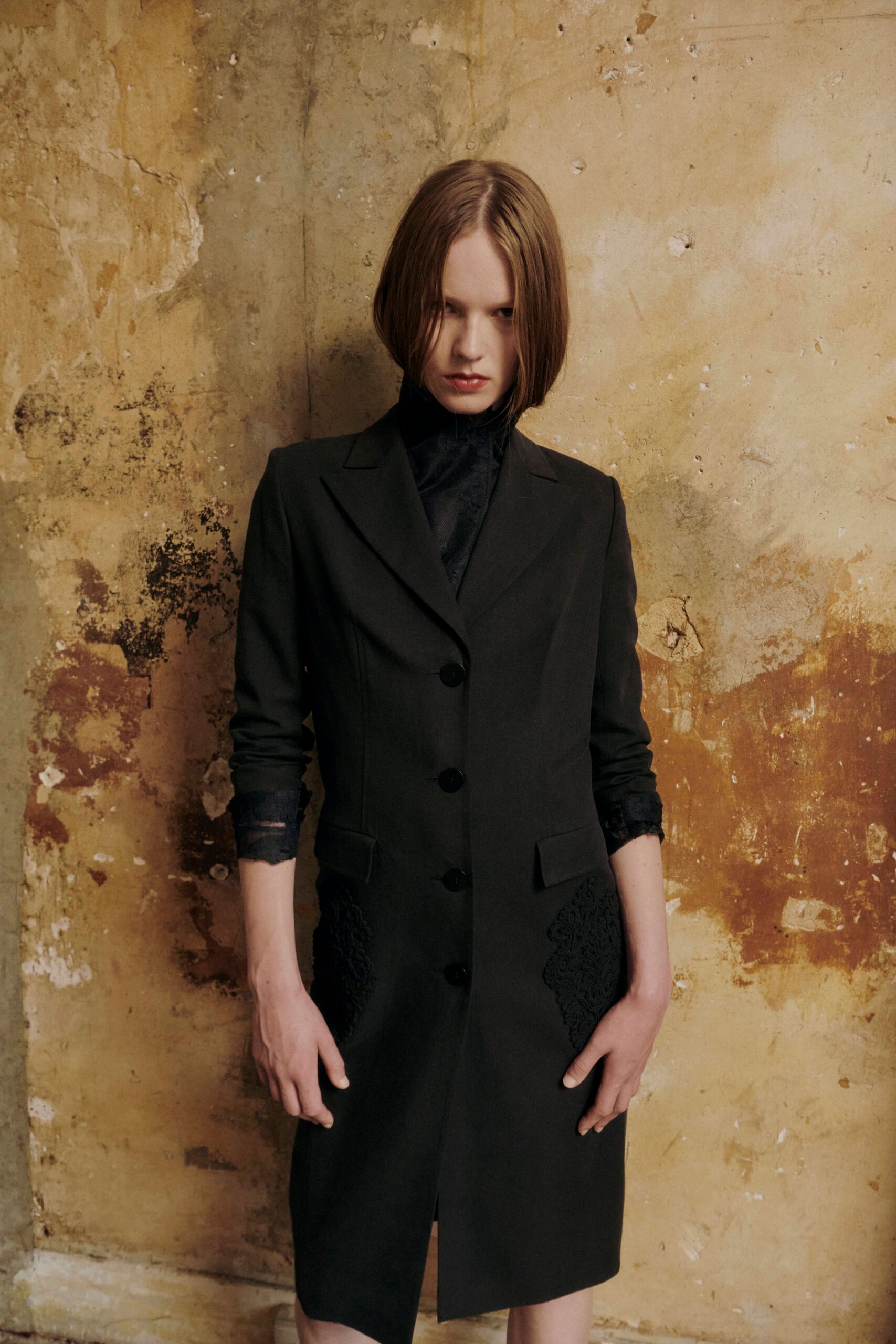 Black tailored coatdress with four covered buttons and raised appliqué at the hips. Sheer black high-neck blouse, lace cuffs just escaping the sleeves. In a crumbling hotel corridor before the radiator heat fully wakes up.