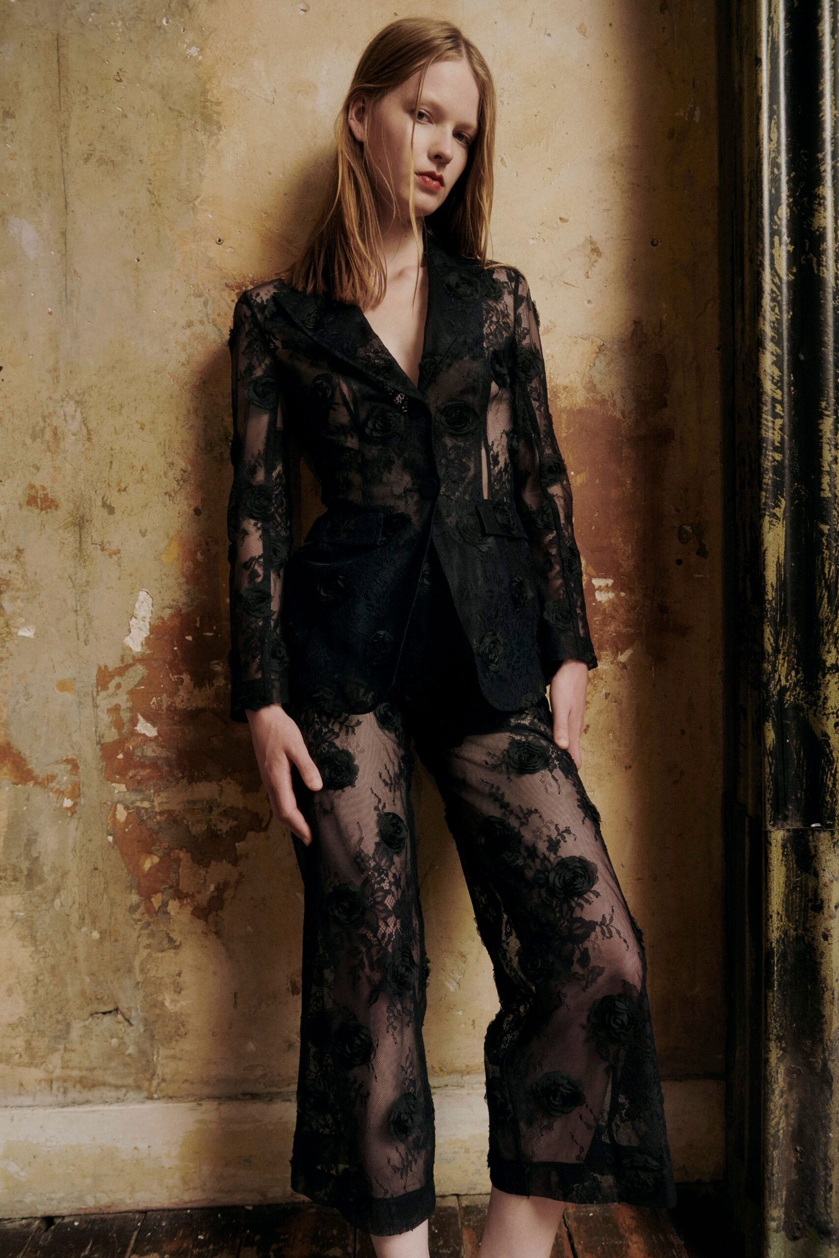 Black blazer in sheer mesh, raised rosettes climbing over the lapels. Matching cropped trousers, floral lace opening to transparent legs above the hem. In a faded hotel corridor before anyone else has come downstairs.