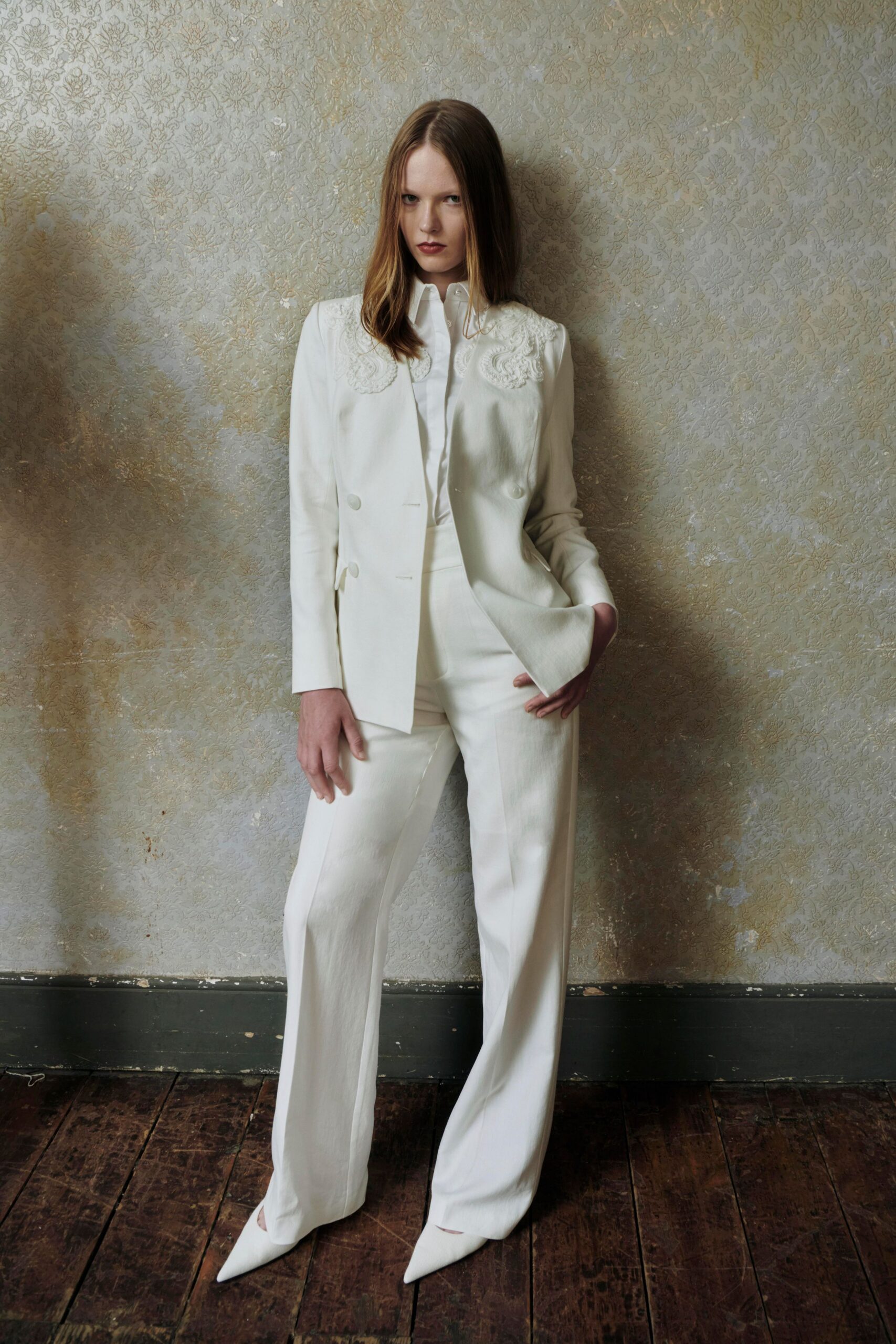 Ivory double-breasted blazer with corded embroidery curling over both shoulders. White shirt buttoned high, collar points standing away from the throat. High-waisted ivory trousers falling in a long fluid break. Sharp white pumps peeking from under the hem. In a faded manor drawing room before anyone opens the piano.