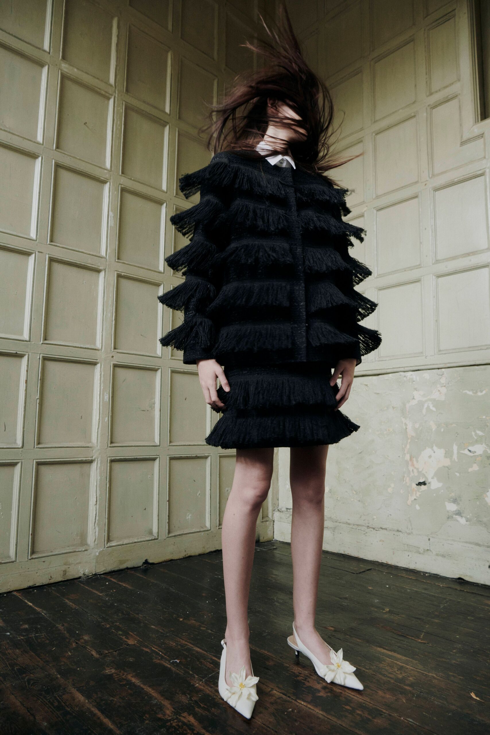 White pointed collar peeking sharp above the neckline. Black jacket built in stacked bands of dense swung fringe. Matching mini skirt with two deep fringe tiers at the hem. White pointed slingbacks topped with oversized fabric flowers. In a drafty manor room, just after someone flung the windows open.